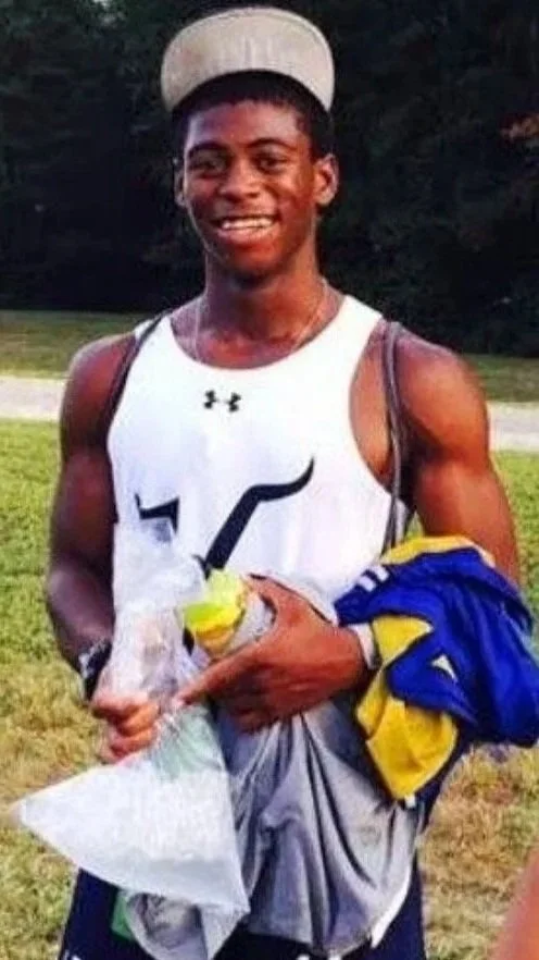 Young man smiling outdoors, wearing a white athletic tank top, a beige hat, and holding sports gear and a plastic bag.
