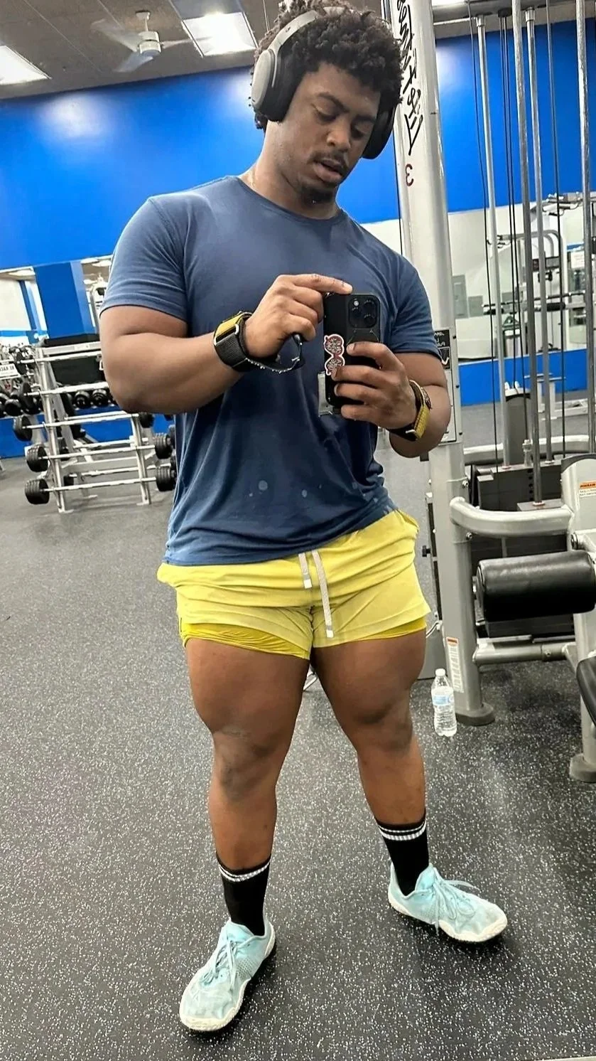 A man at a gym taking a mirror selfie, wearing a blue T-shirt, yellow shorts, black socks, and light blue sneakers, with headphones and a wristwatch.