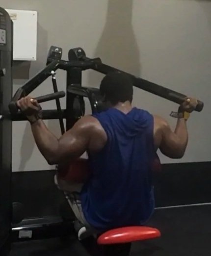 A person with a muscular back working out at the gym, sitting on a chair while holding a barbell on their shoulders.