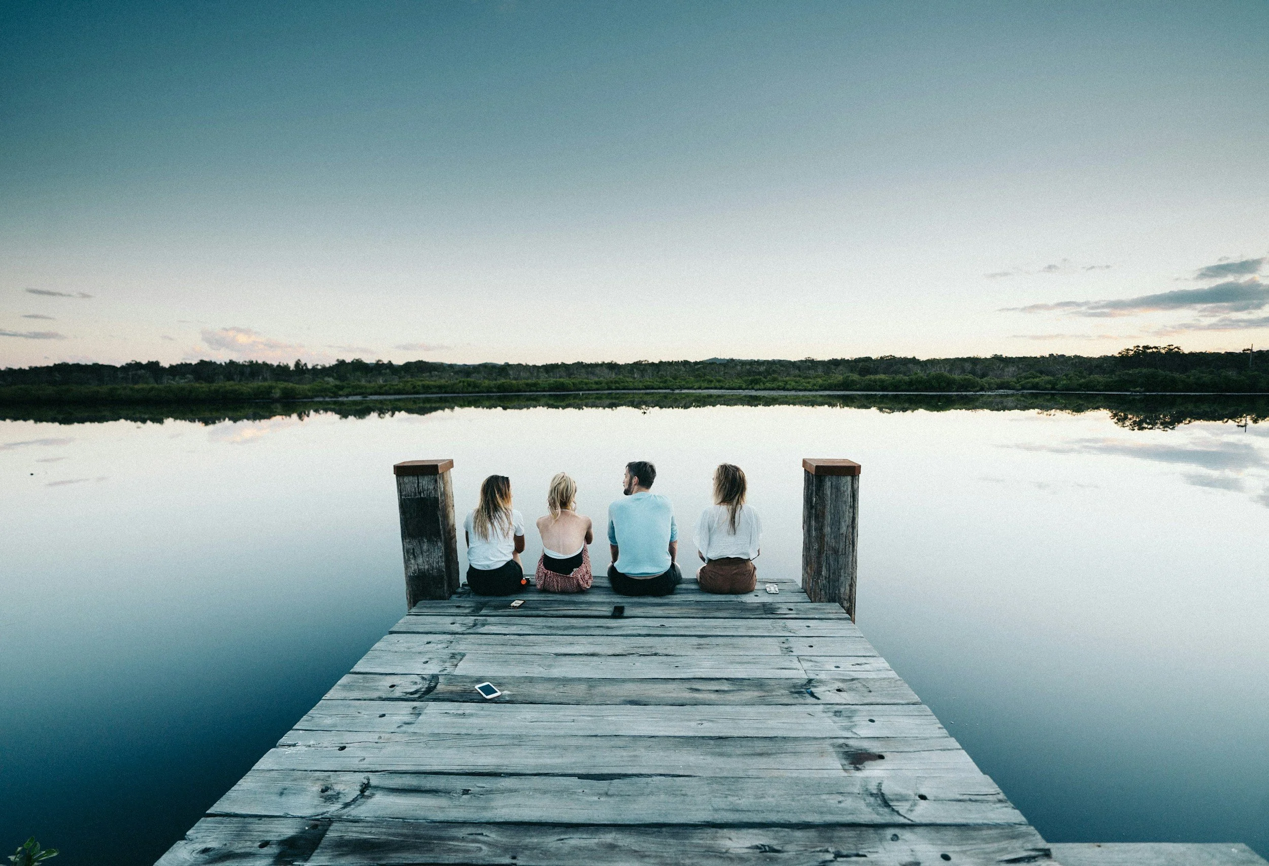 Four people sitting on a wooden dock by a calm lake at dusk, looking at the water and distant shoreline with trees.