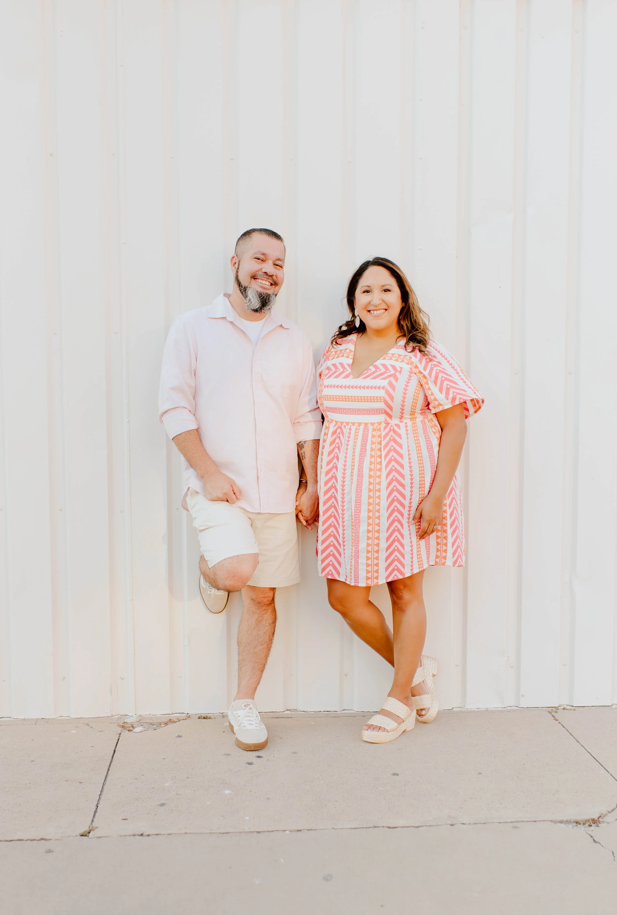 A man with a beard wearing a light pink shirt and beige shorts standing next to a woman in a pink and white patterned dress, both smiling and holding hands against a white wall.