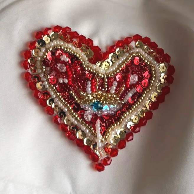Decorative heart-shaped art piece made of red, gold, white, and blue beads on a white fabric background.