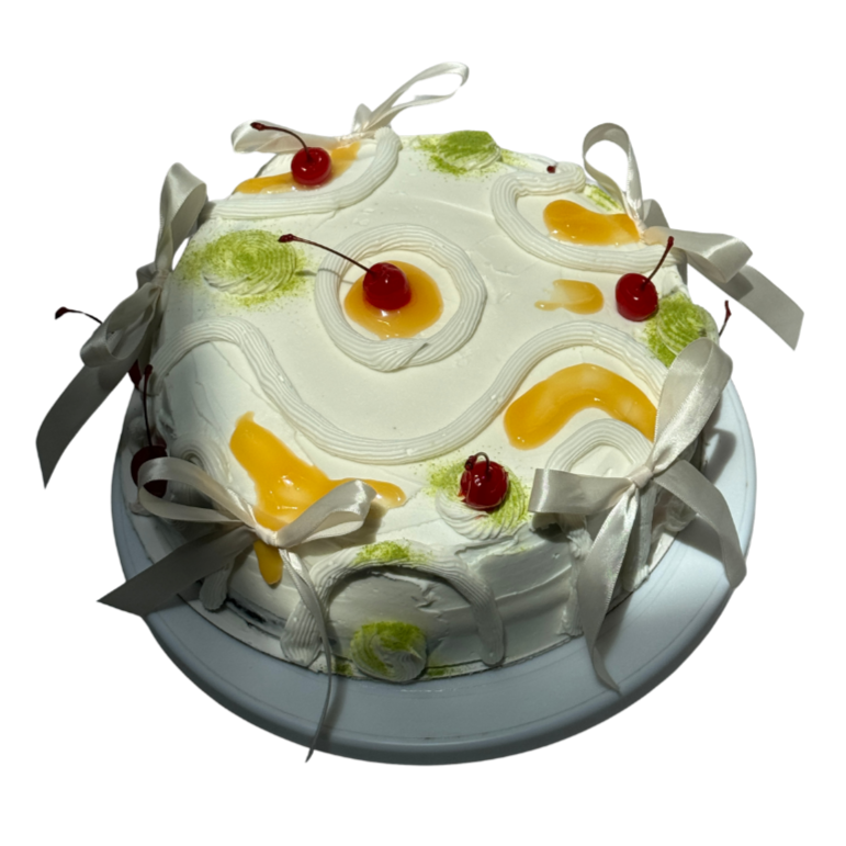Decorated round cake with white frosting, cherries on top, yellow and green icing accents, and white ribbons around the edges.