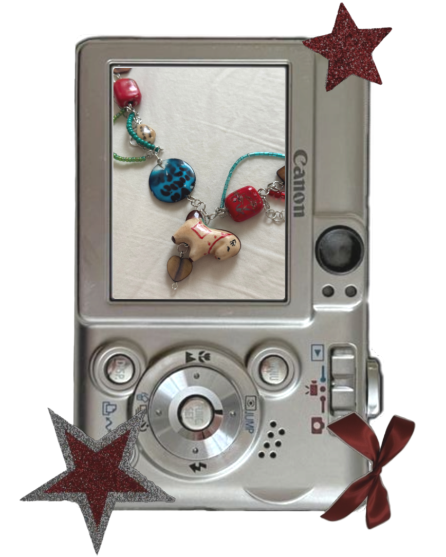 A digital camera with a silver body and a black control button displays a photo of a necklace with colorful beads and charms, resting on a white surface. The camera is decorated with red and silver glittery stars and a red bow in the corners.