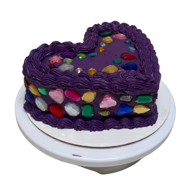 A heart-shaped birthday cake decorated with purple frosting and colorful gemstones on top and sides, placed on a white cake stand.