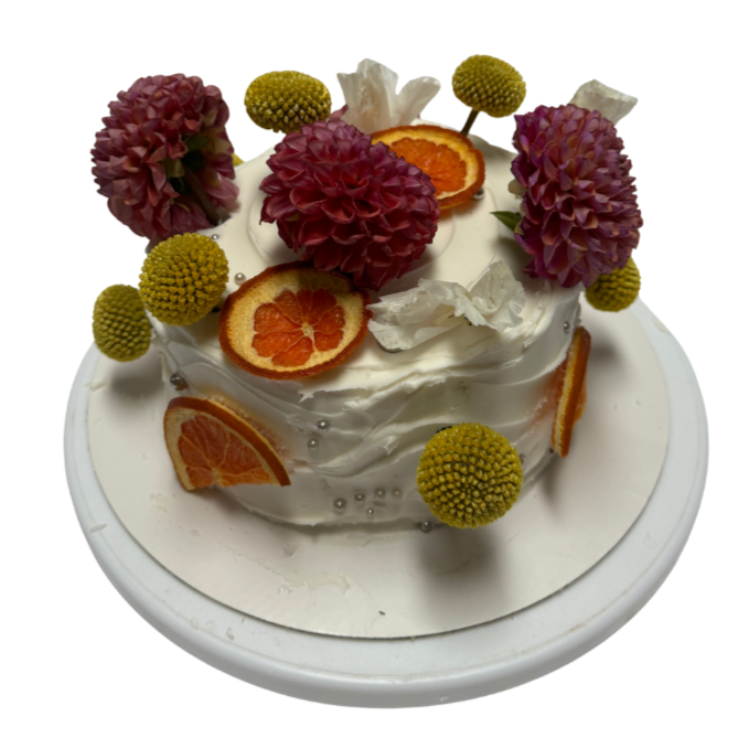 Decorated cake with white frosting, pink flowers, orange slices, white flowers, and green spherical accents on a white round cake board.