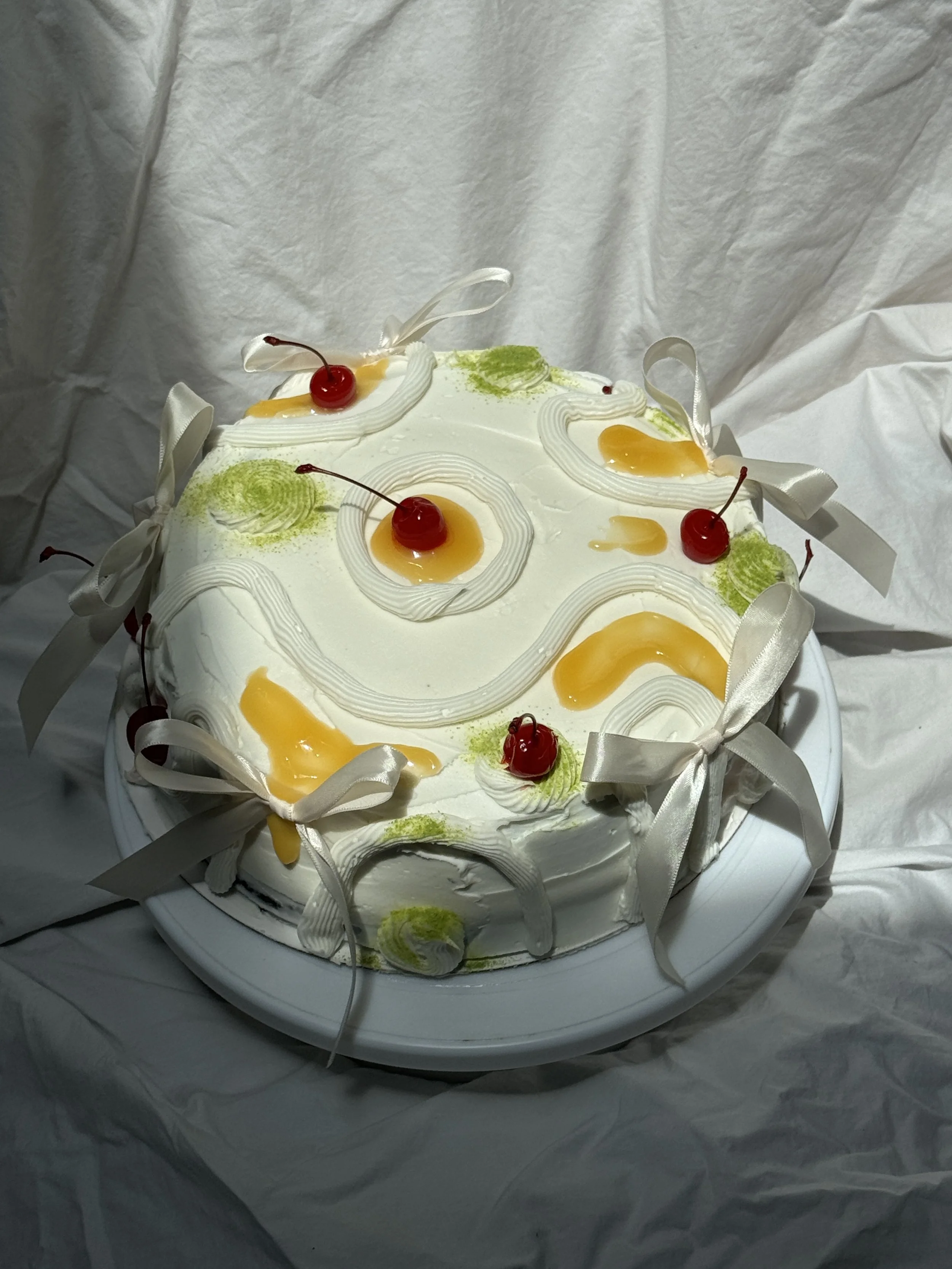 A round white cake decorated with cherries, cream, green and yellow icing, and white ribbons, sitting on a white cake stand.