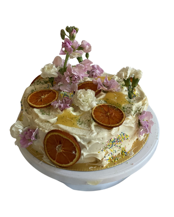 A decorated cake with white frosting, dried orange slices, pink and white flowers, rainbow sprinkles, and a drizzle of honey.
