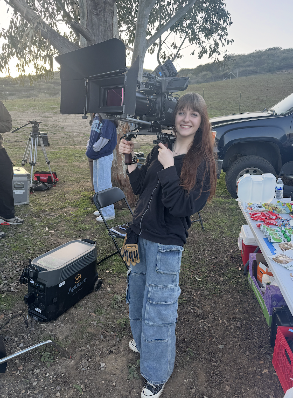 A young woman with long brown hair, wearing a black hoodie, loose jeans, and sneakers, is smiling and holding a large professional camera on a stabilizer in an outdoor setting. There are film crew members, equipment, and a table with snacks nearby, with a truck and a tree in the background.