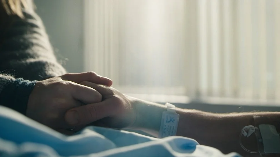 Person in a hospital bed holding hands with someone, wearing an IV line.
