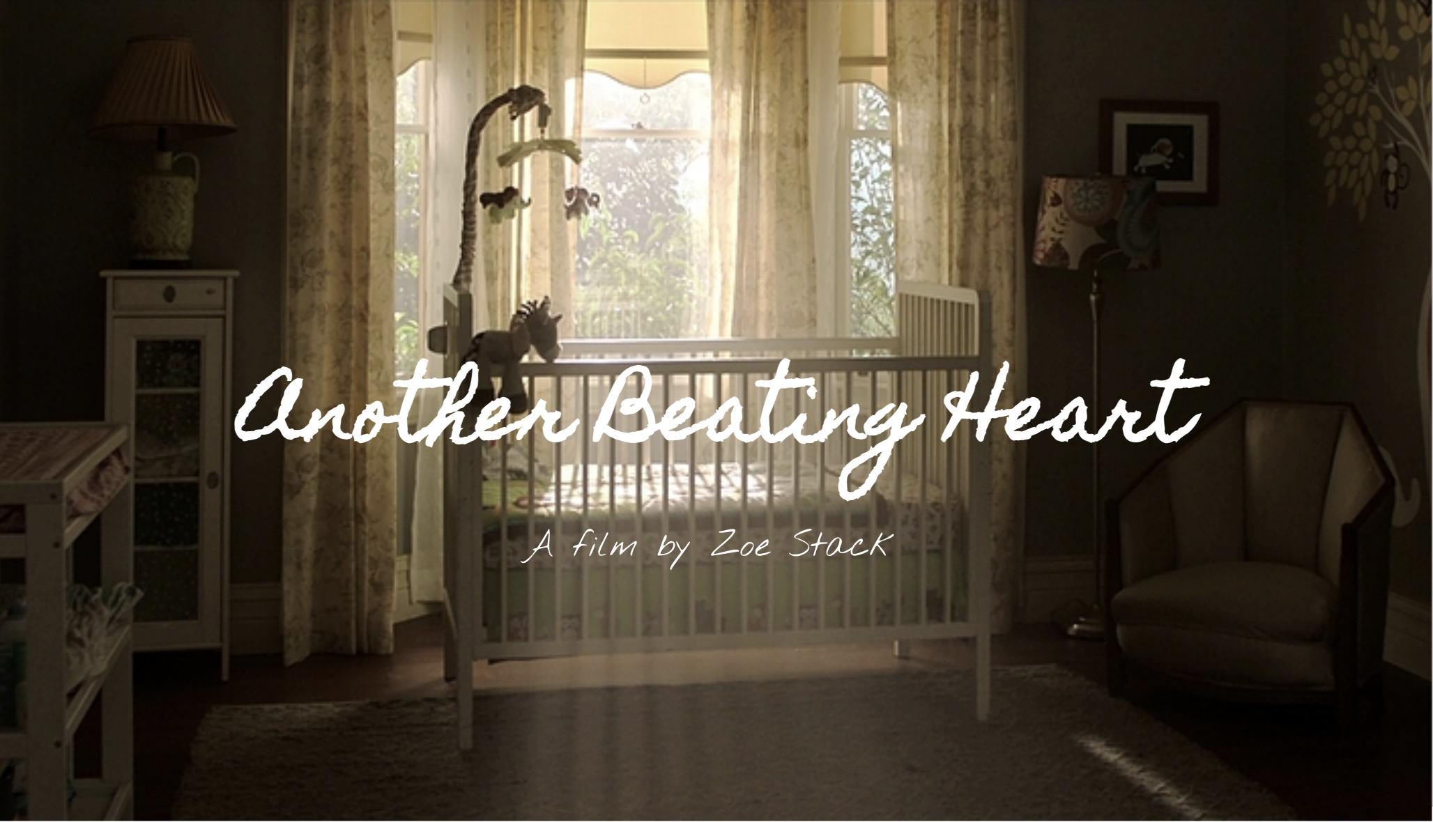 A dimly lit nursery with a white crib in front of a large window with floral curtains, a giraffe-shaped mobile hanging above the crib, and a plush zebra toy inside. There is a white cabinet with a lamp on top on the left and a beige armchair with a c