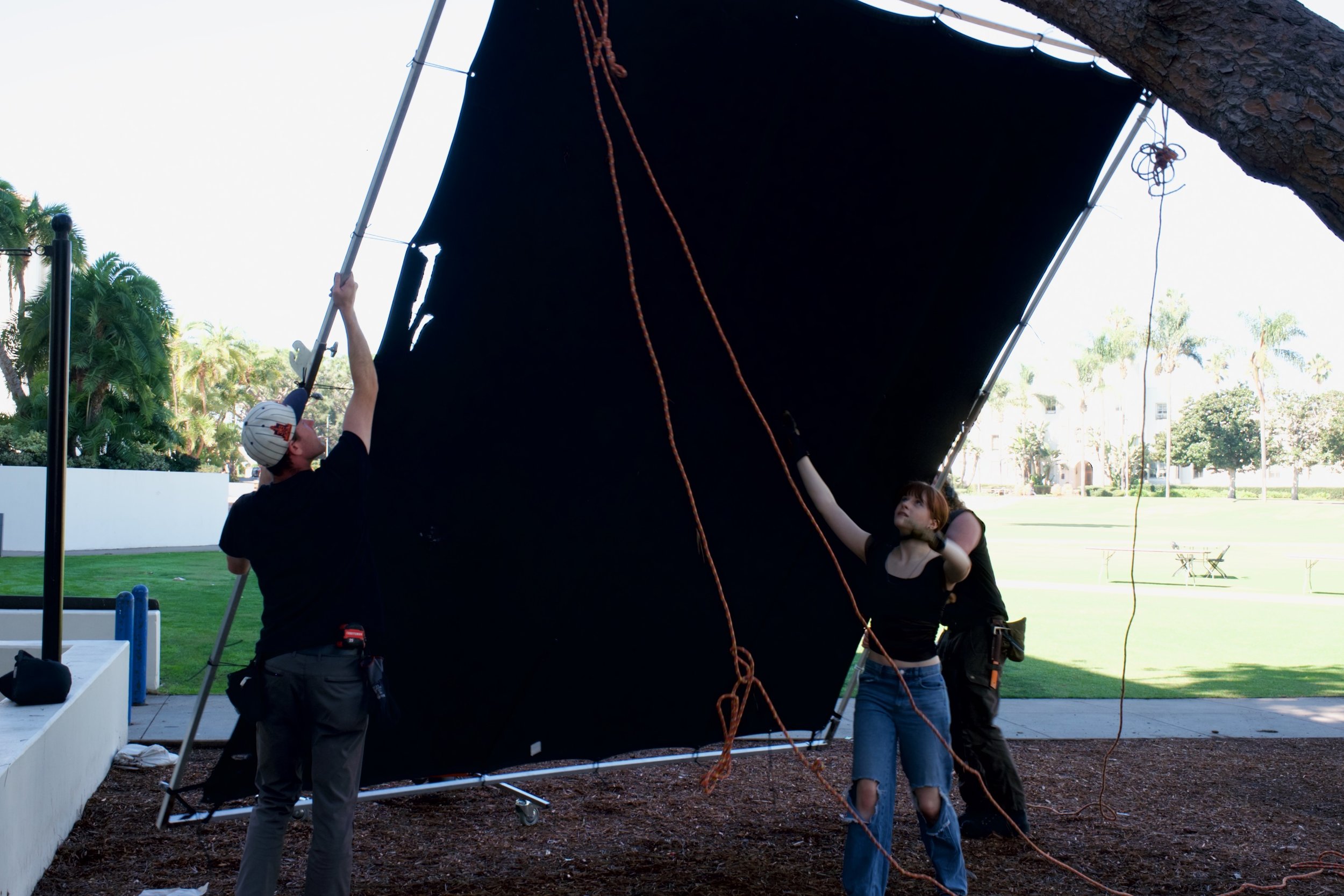 Three crew members work together on a film set, adjusting a large black screen outdoors on a sunny day, with trees and a park in the background.
