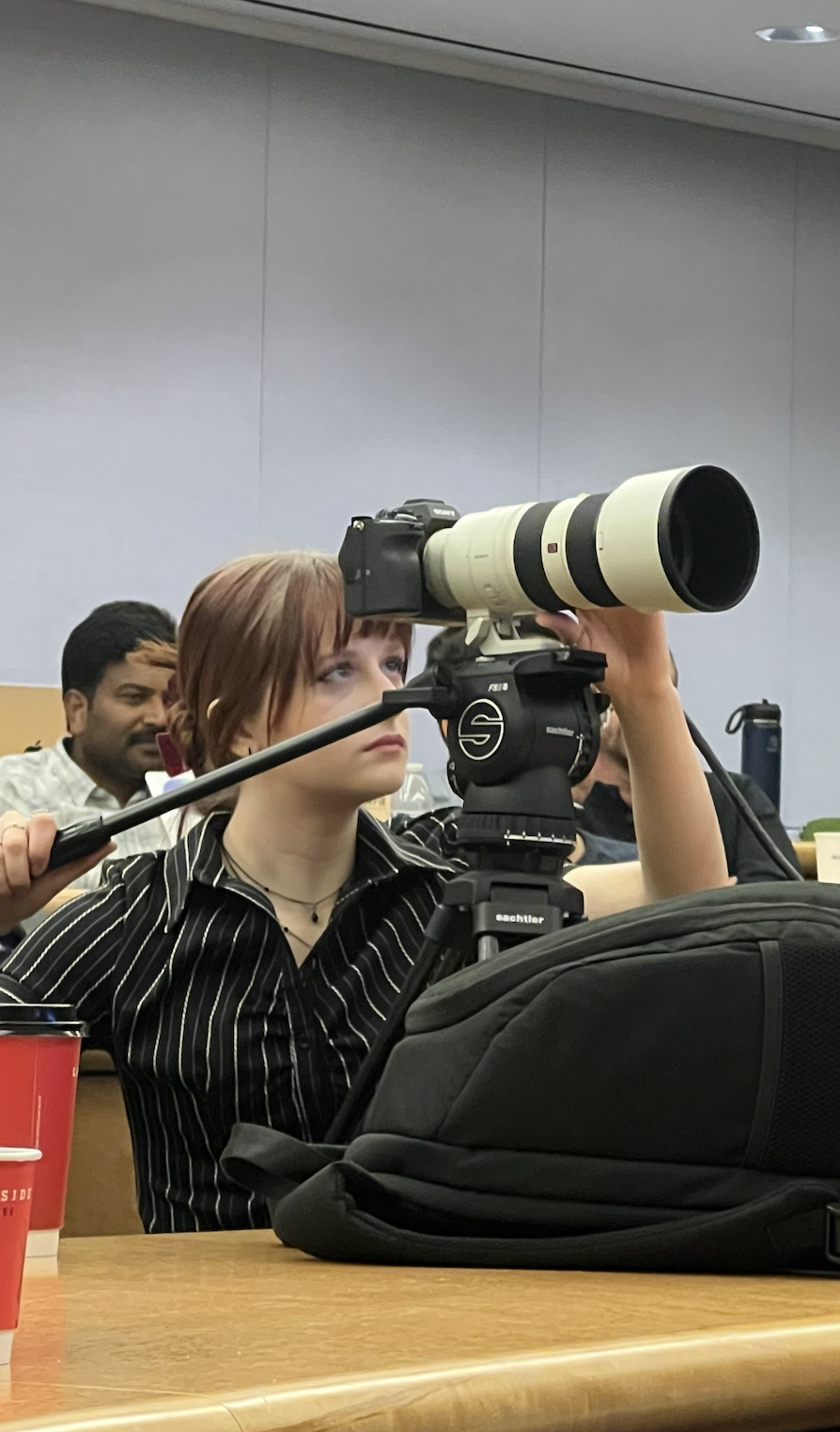 A young woman with short red hair using a large telephoto camera lens at a conference or classroom setting. She is seated at a table with a black bag and red cups nearby, focused on her photography.
