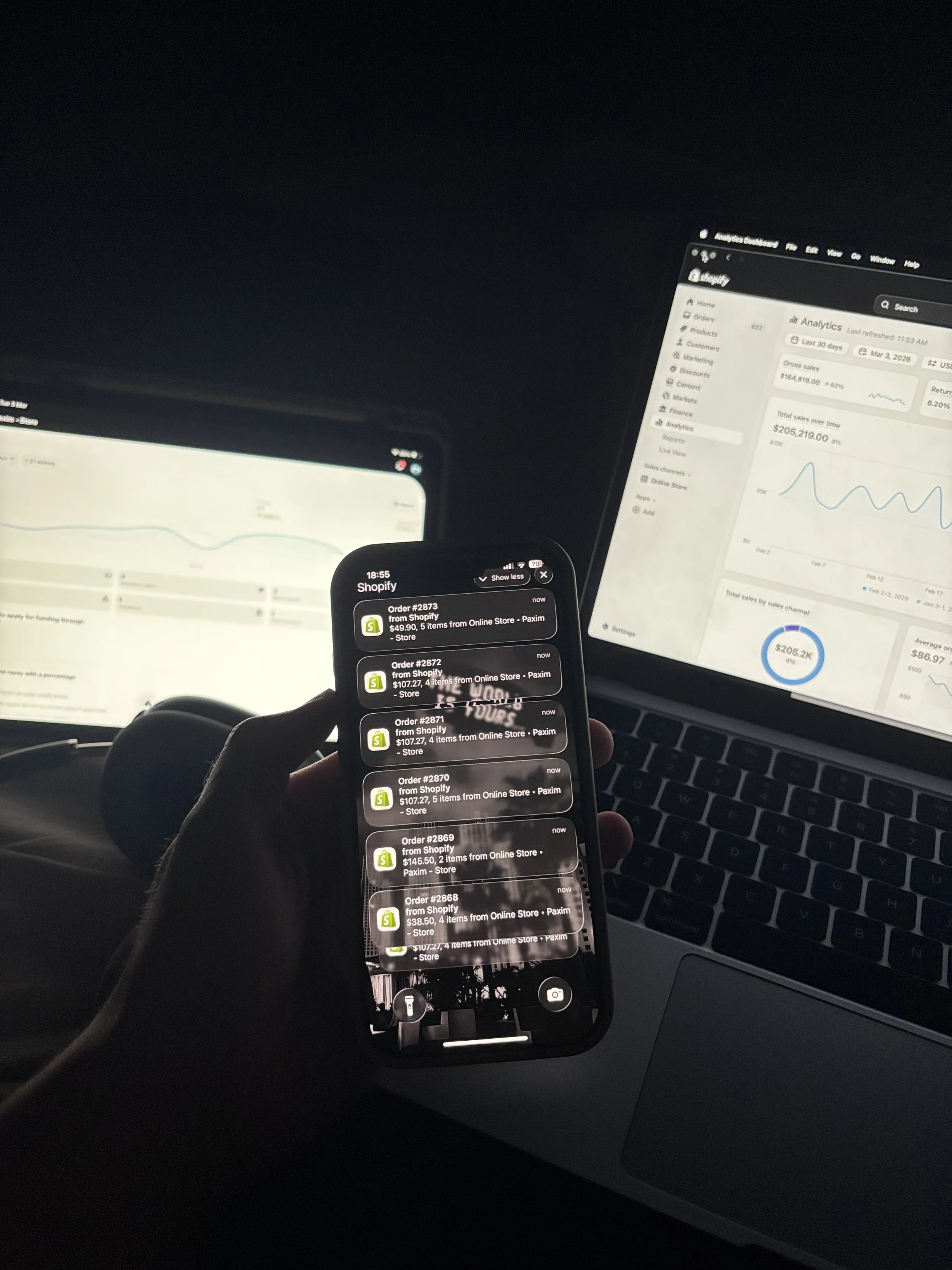 Person holding a smartphone displaying Shopify order notifications, with laptop screens in the background showing analytics and sales data.