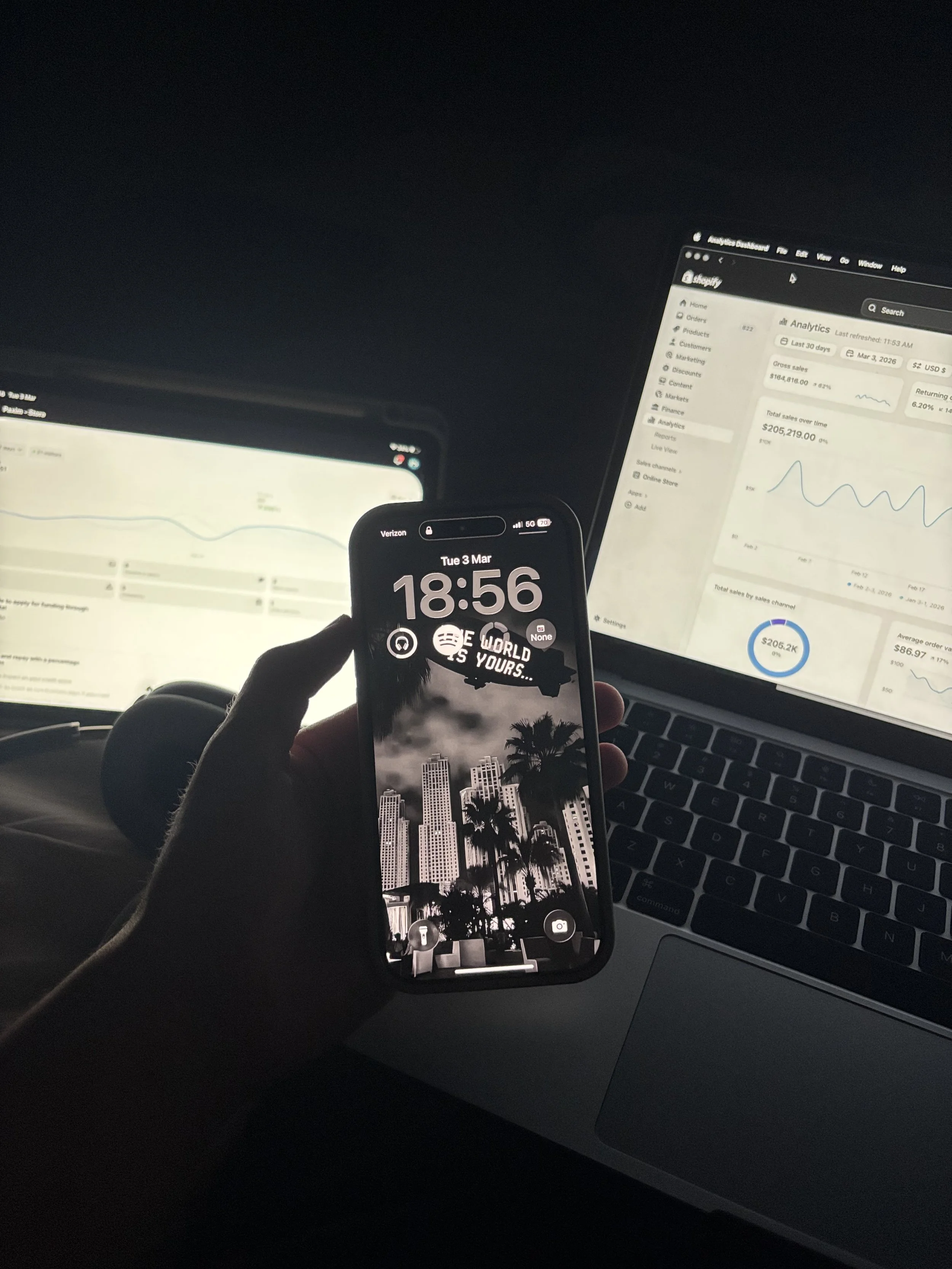 Photo taken inside a car showing a person holding a smartphone with an image of a cityscape with palm trees in the foreground. The phone's screen displays the time as 18:56 on Tuesday, March 3. In the background, there are laptop screens showing analytics dashboards with graphs and data.