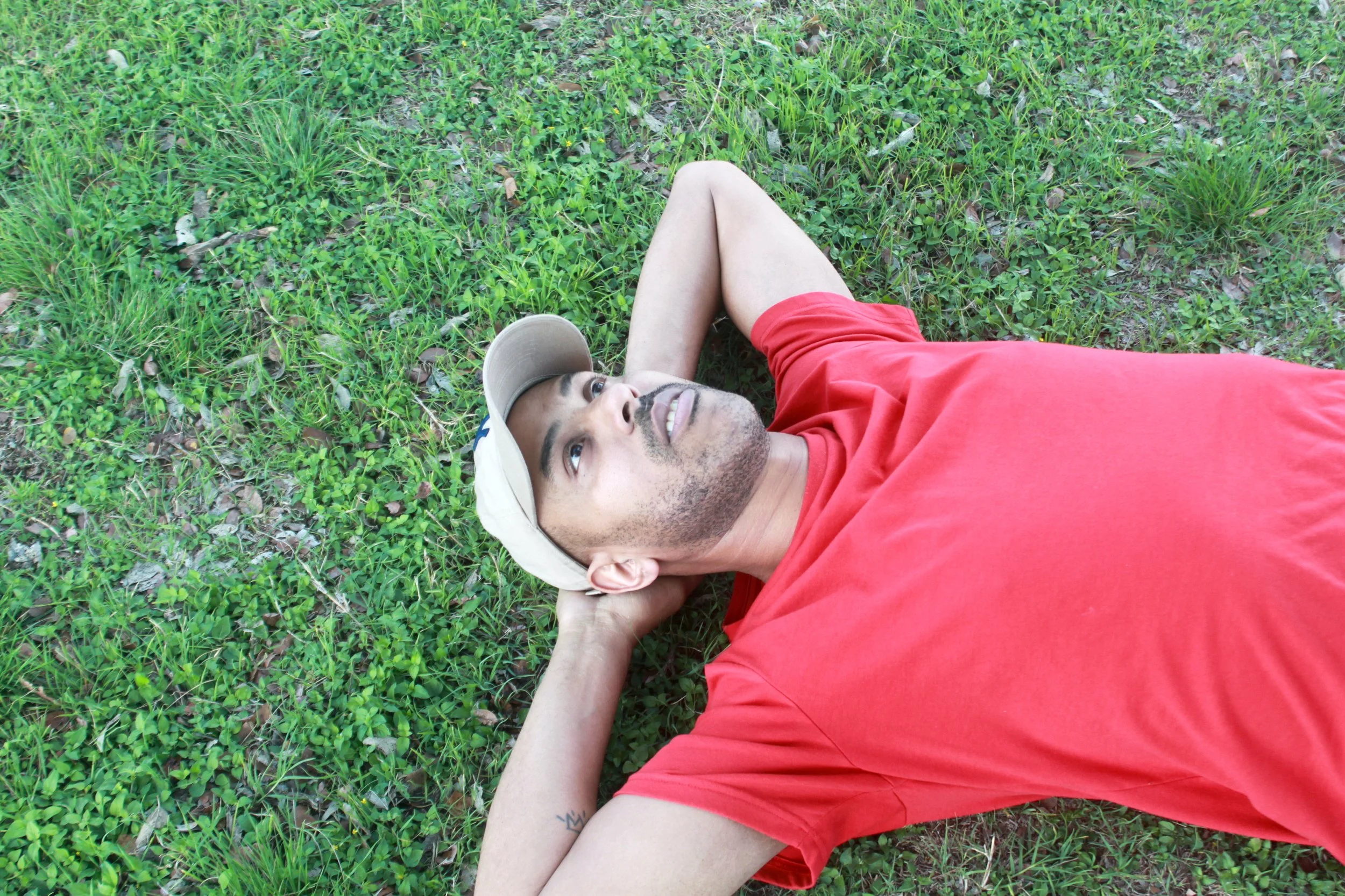 A man lying on his back on green grass, wearing a red t-shirt and a beige cap, looking up with a relaxed expression.