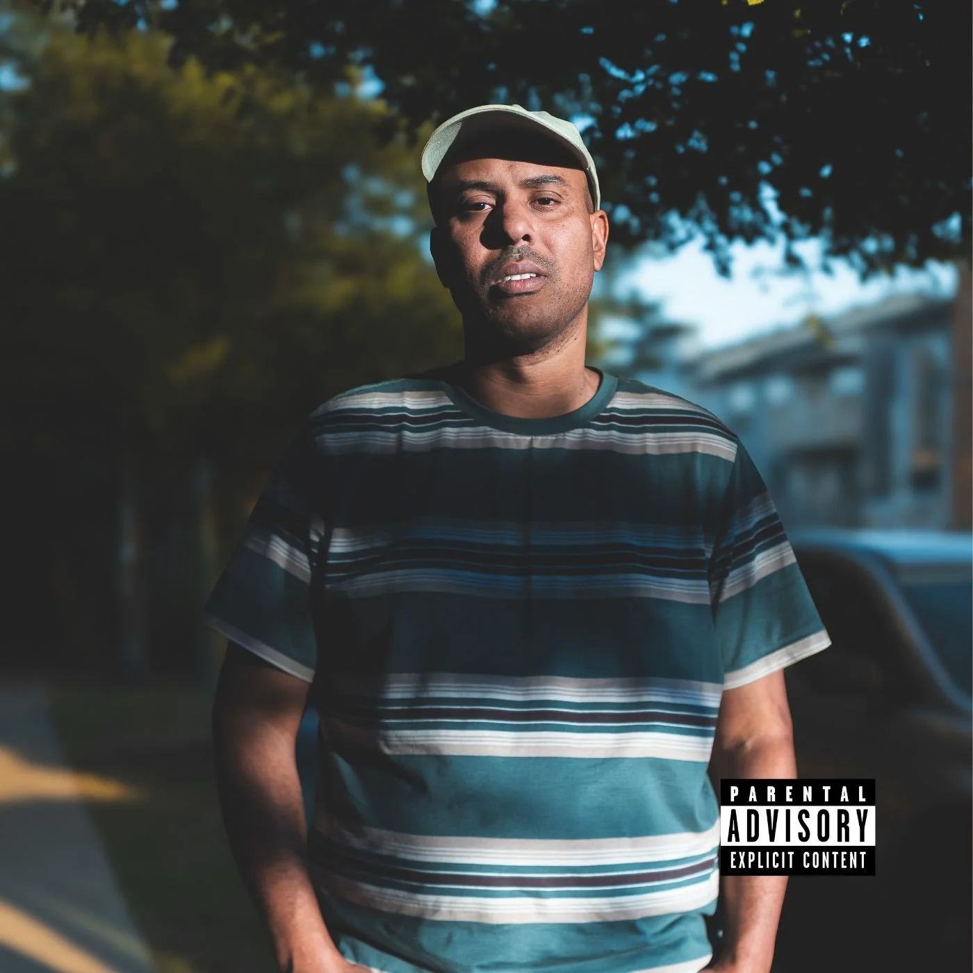 A man wearing a striped T-shirt and a white cap stands outdoors during the daytime, with trees and a building visible in the background, and a "Parental Advisory" warning label in the bottom right corner.