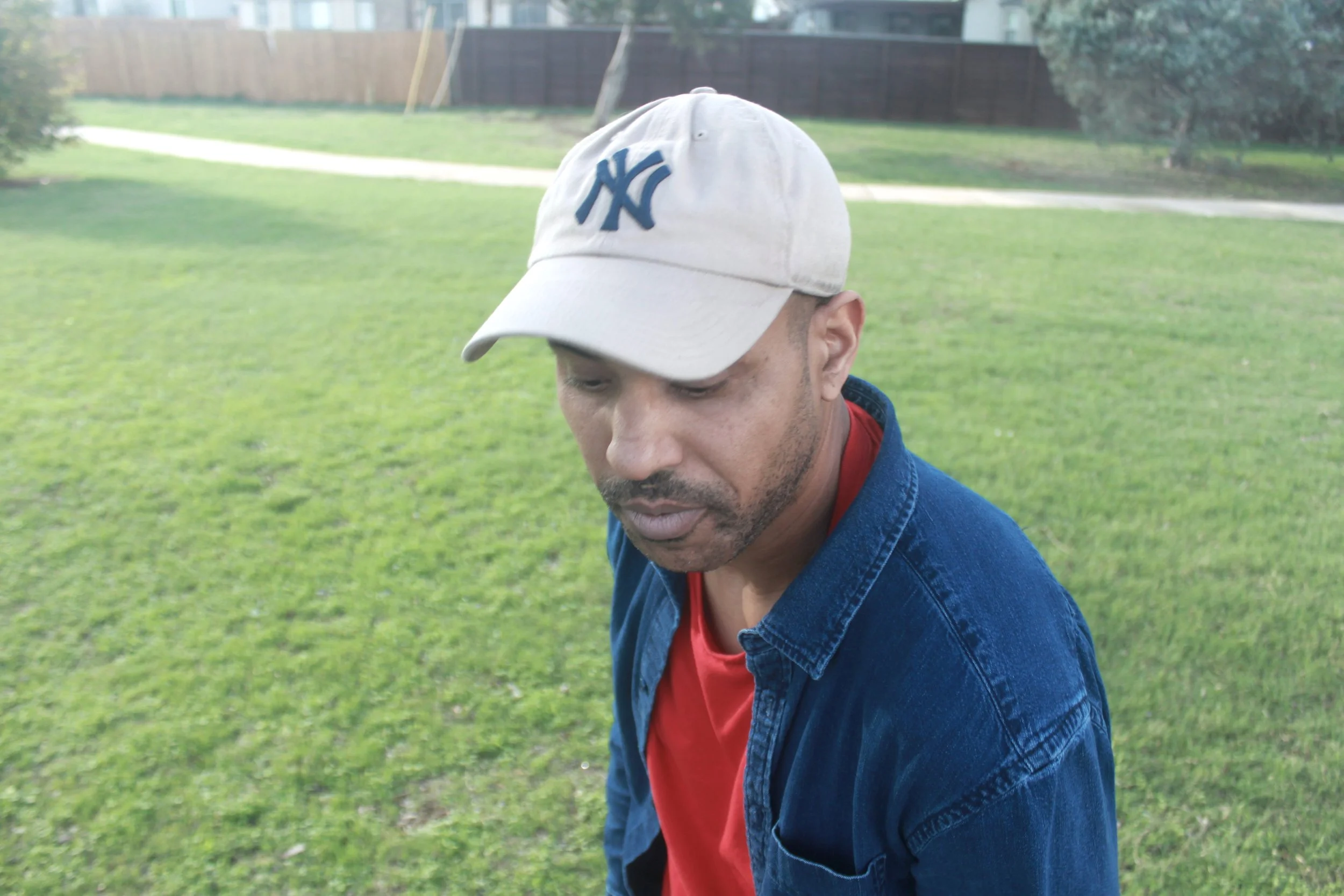 A man wearing a white New York Yankees baseball cap, a red shirt, and a denim jacket, looking down and standing on a grassy field in an outdoor park.