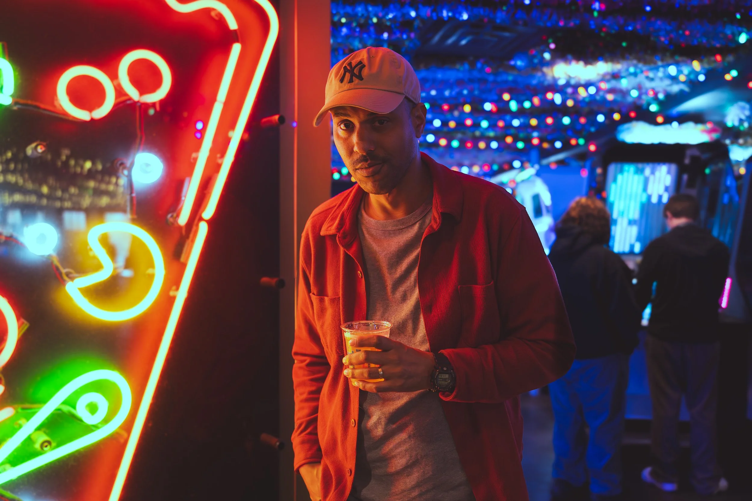 A man in a beige cap and red jacket holding a drink stands near a bright neon pinball machine at night, with colorful string lights and other people in the background.