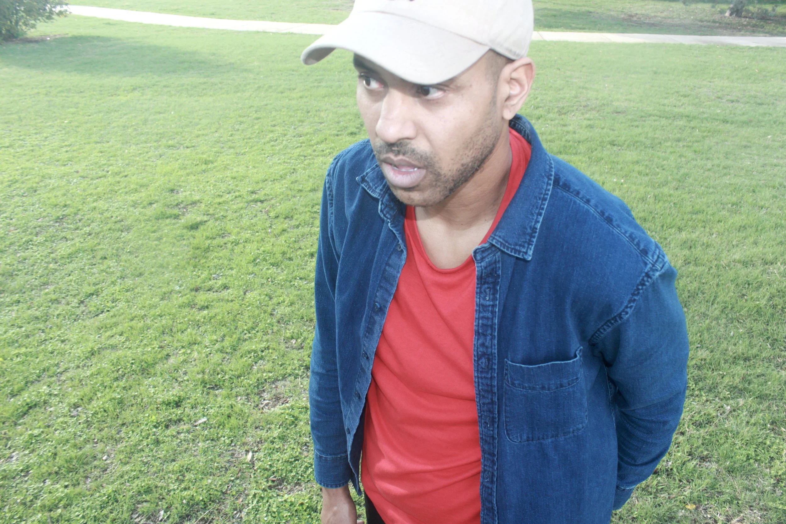 A man wearing a beige baseball cap, a red shirt, and a blue denim jacket standing on a green grassy field, looking to the side with a surprised or curious expression.