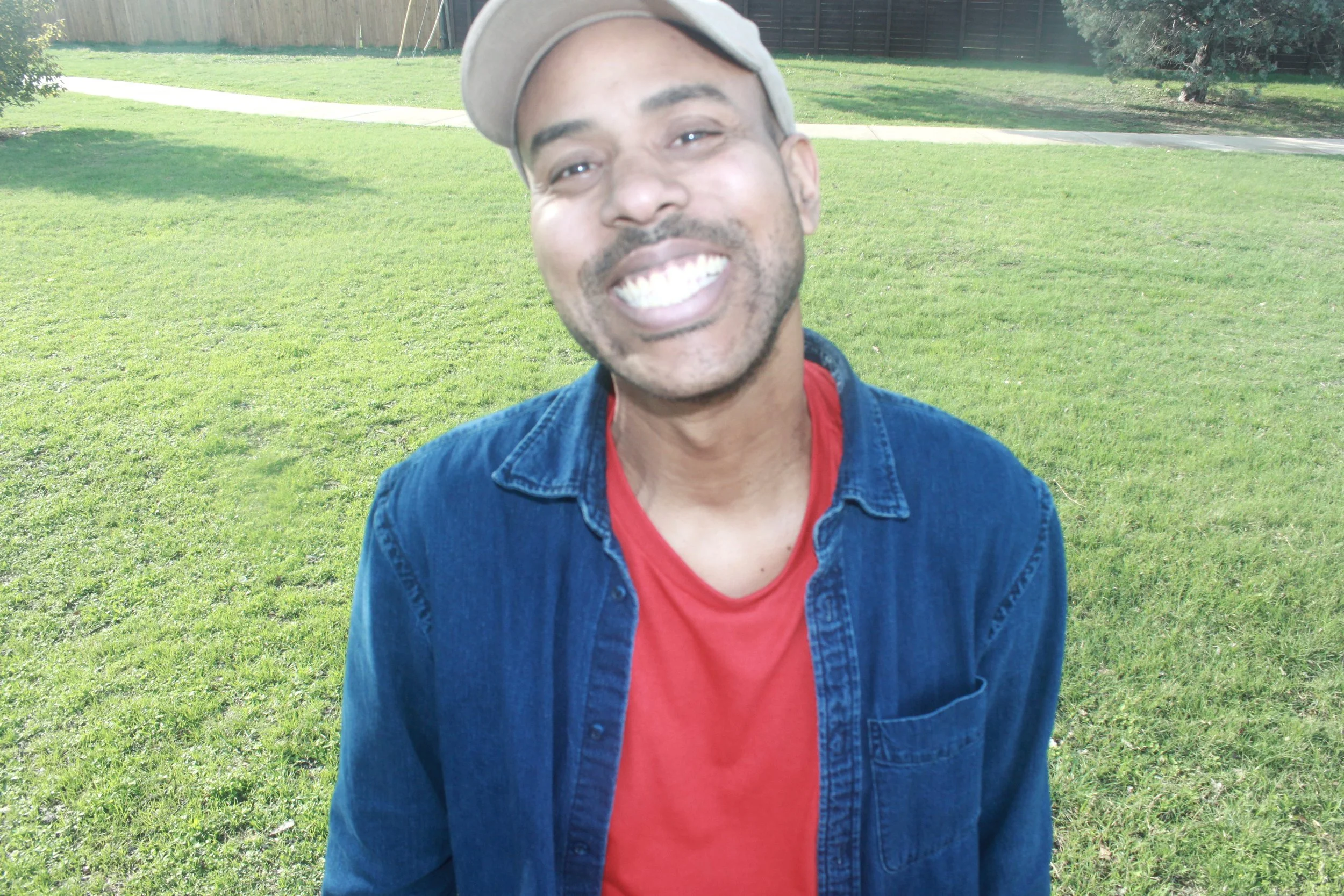 A man smiling outdoors on a grassy field, wearing a beige cap, red t-shirt, and denim jacket.