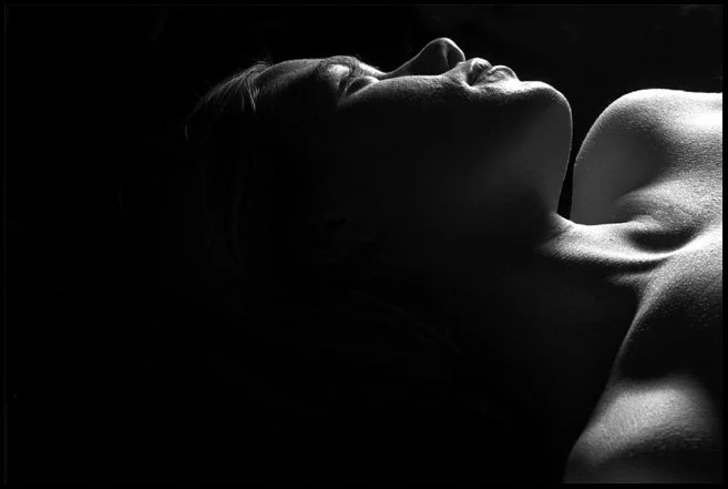 A person lying on their side with their eyes closed, partially illuminated with high contrast lighting.