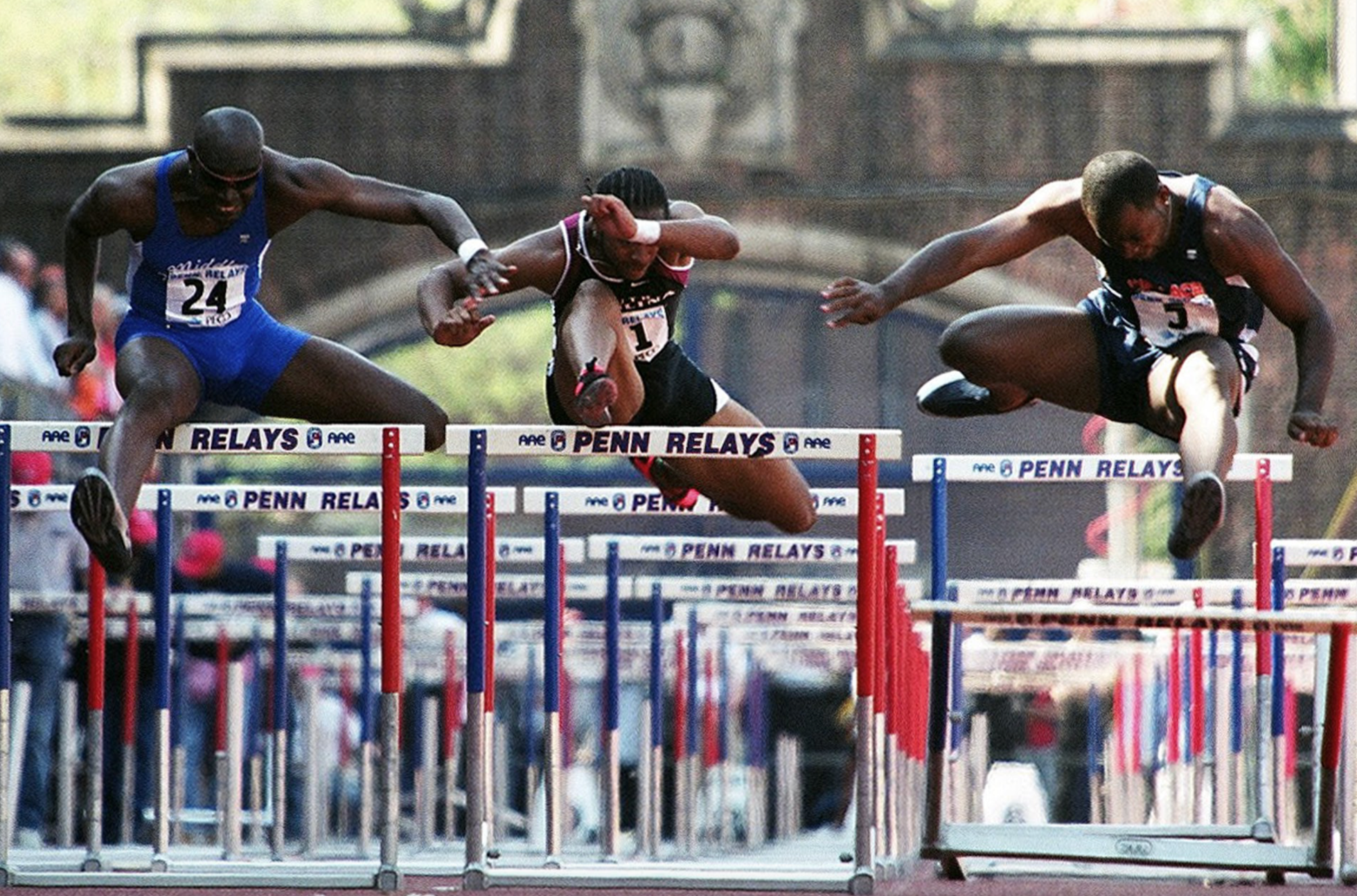 Three athletes competing in a hurdles race at Penn Relays, jumping over hurdles with a crowd in the background.