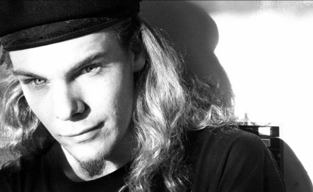 Black and white portrait of a young man with long wavy hair, a goatee, wearing a hat, and looking into the camera.