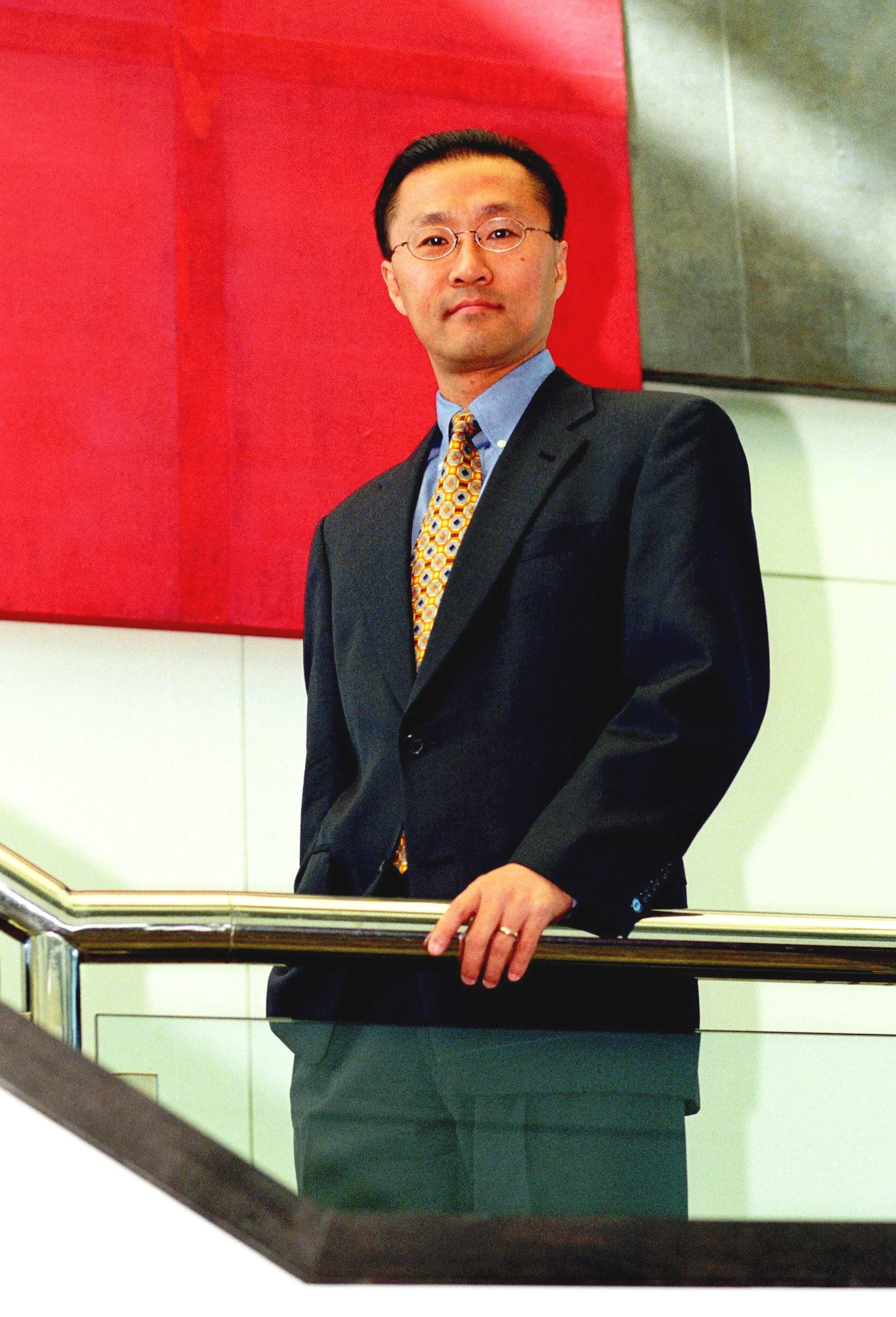 Asian professional man in a dark suit standing in front of a red and gray wall, with a hand resting on a railing.