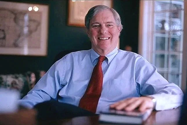 A smiling middle-aged lawyer with gray hair, wearing a light blue dress shirt and red tie, sitting at a desk with a book in front of him in a cozy, well-lit room.