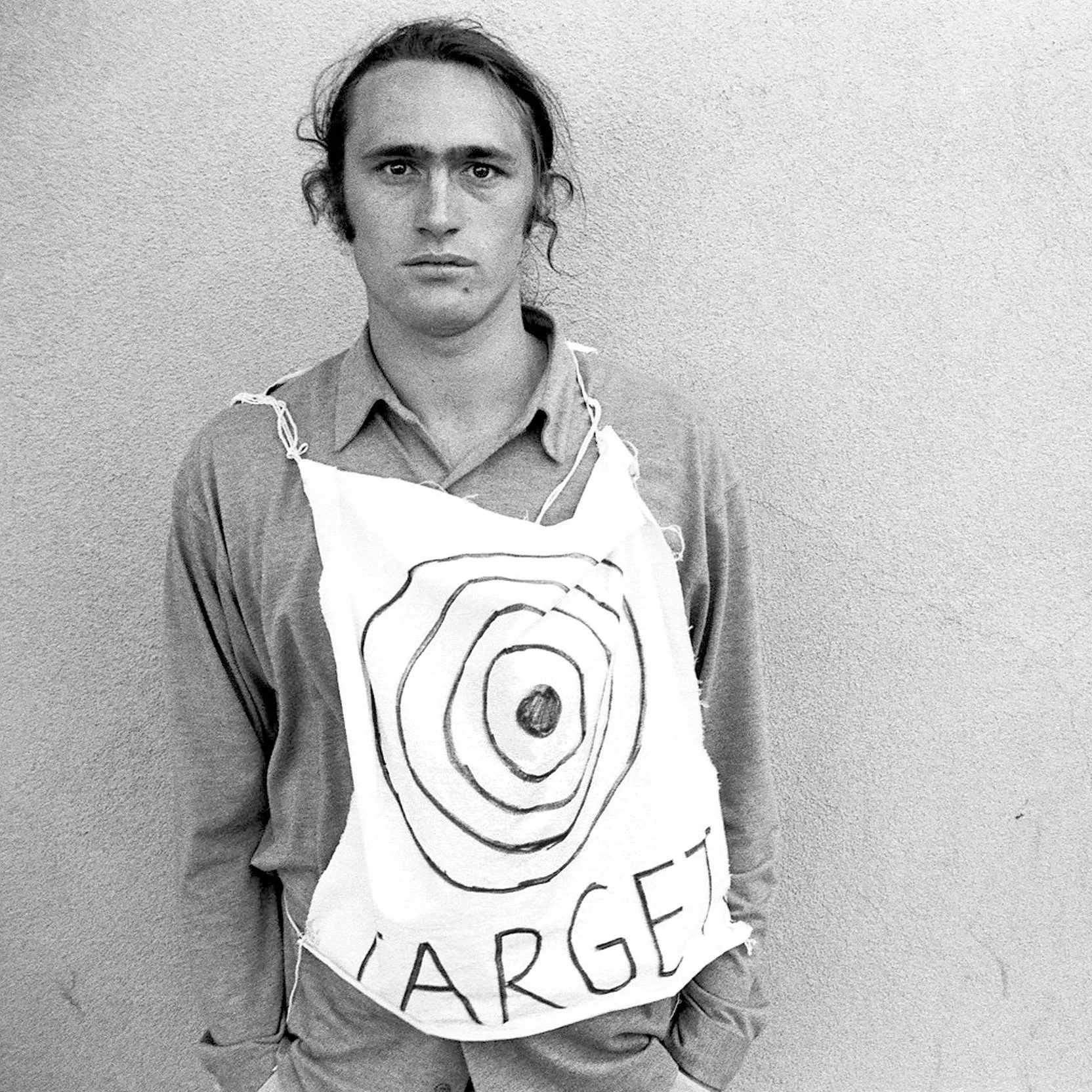 Black and white photo of a person with shoulder-length wavy hair, wearing a shirt and a vest. They are standing against a plain wall and holding a chalkboard with a concentric circle target and the words 'TARGET' written below.