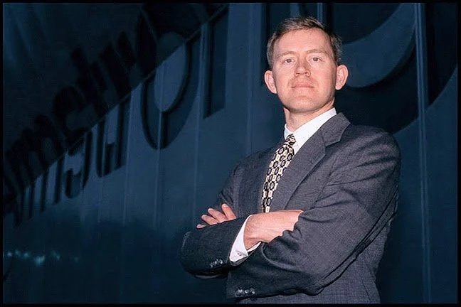 A man in a suit standing outdoors with crossed arms, in front of a building with large illuminated letters.