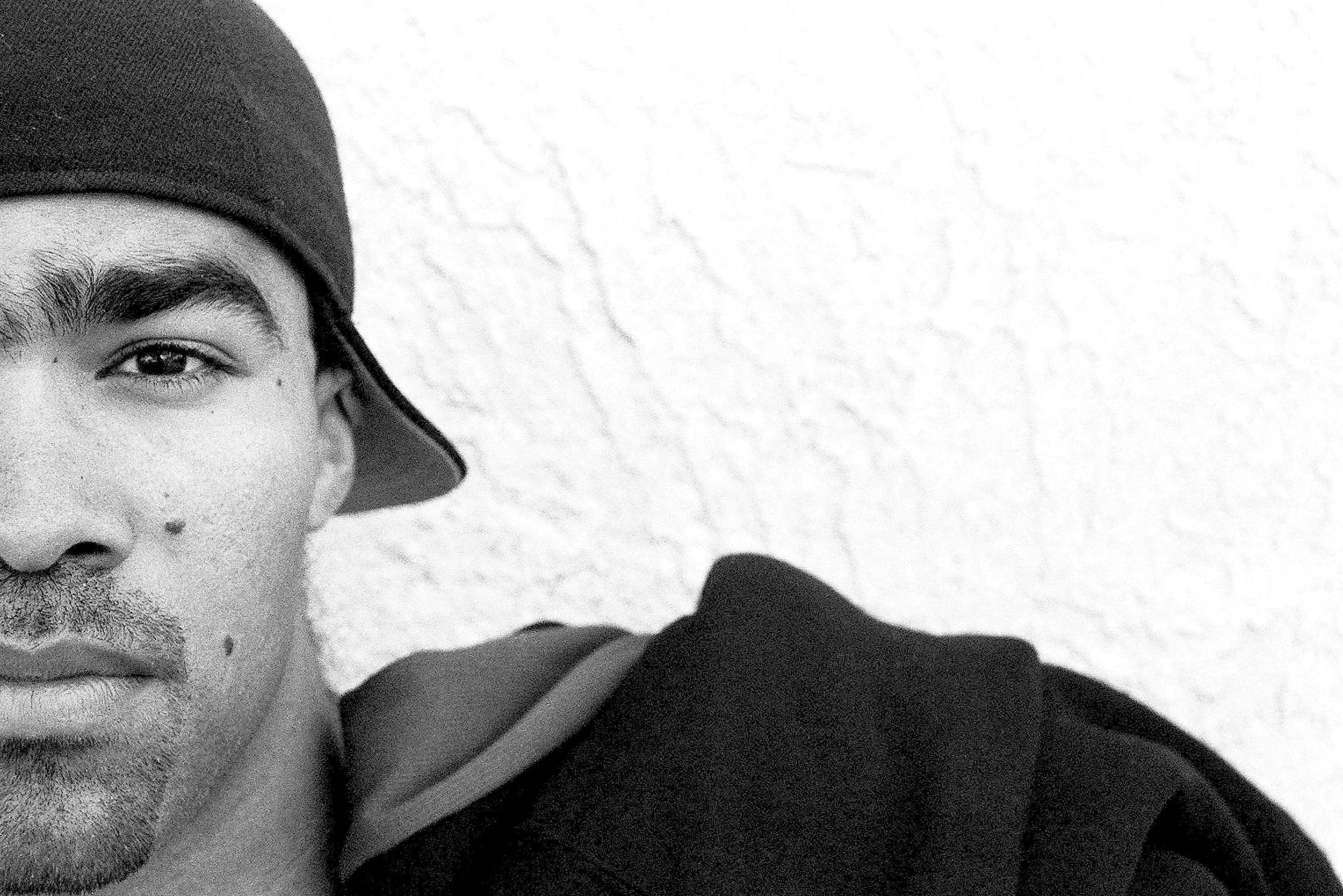 A close-up black-and-white photo of a man wearing a hat, with part of his face, eye, nose, and mouth visible, and a person's hand partially covering his face.