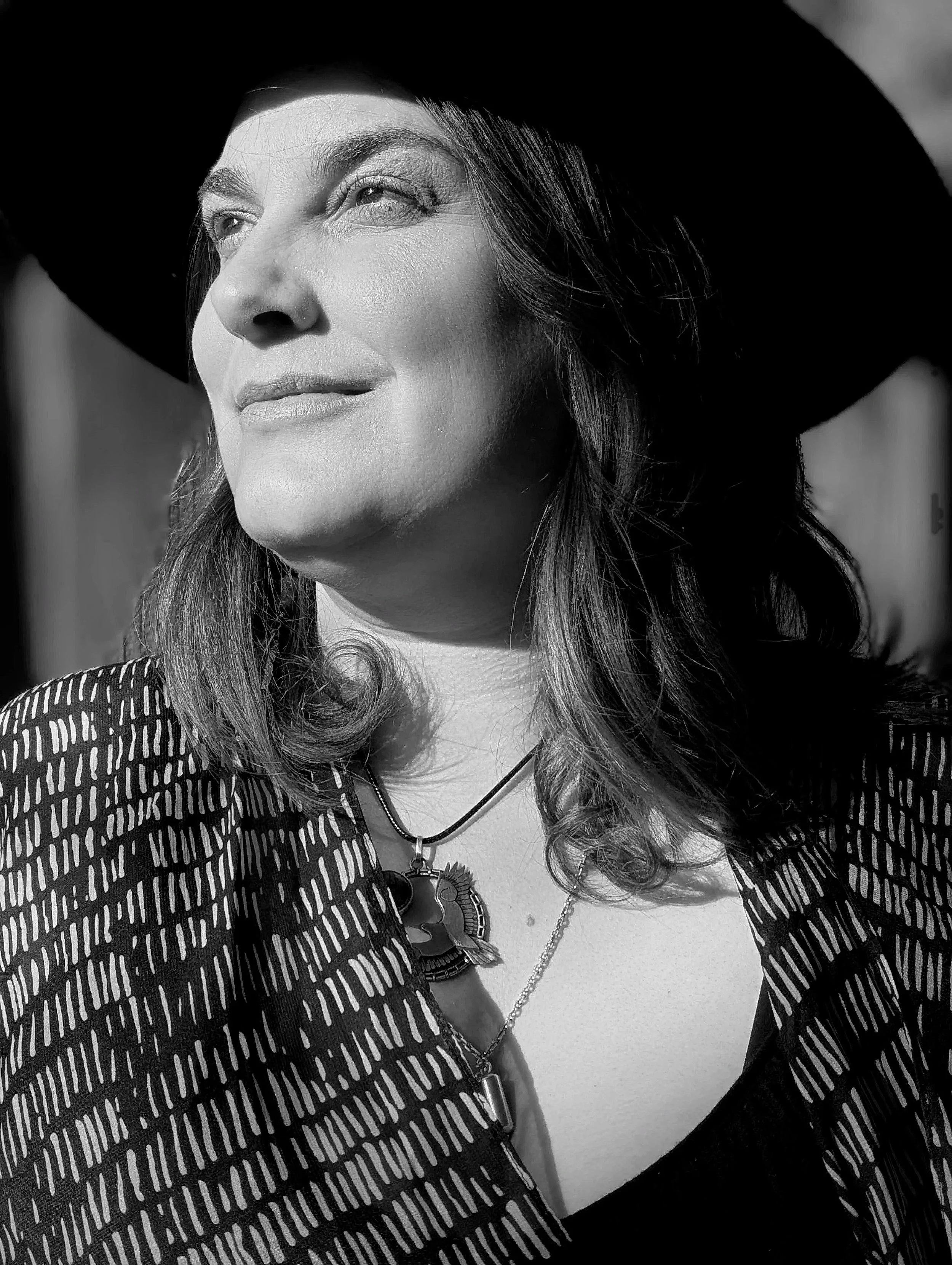 A professional woman in black and white wearing a large black hat and a patterned dress looking towards the sun, with a necklace and a pendant