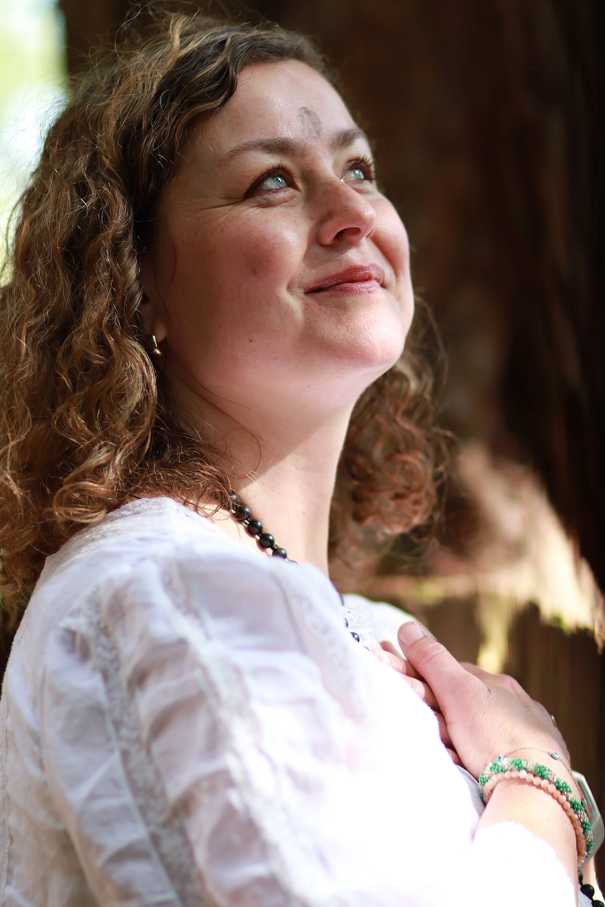 Close-up of a woman with curly brown hair, blue eyes, wearing a white blouse, beaded necklace, and colorful bracelets, looking upward with a serene expression.