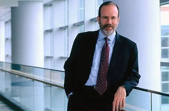 A corporate professional male with glasses and a beard wearing a dark suit, light blue shirt, and patterned tie, standing in a modern office building with large windows and a glass railing.
