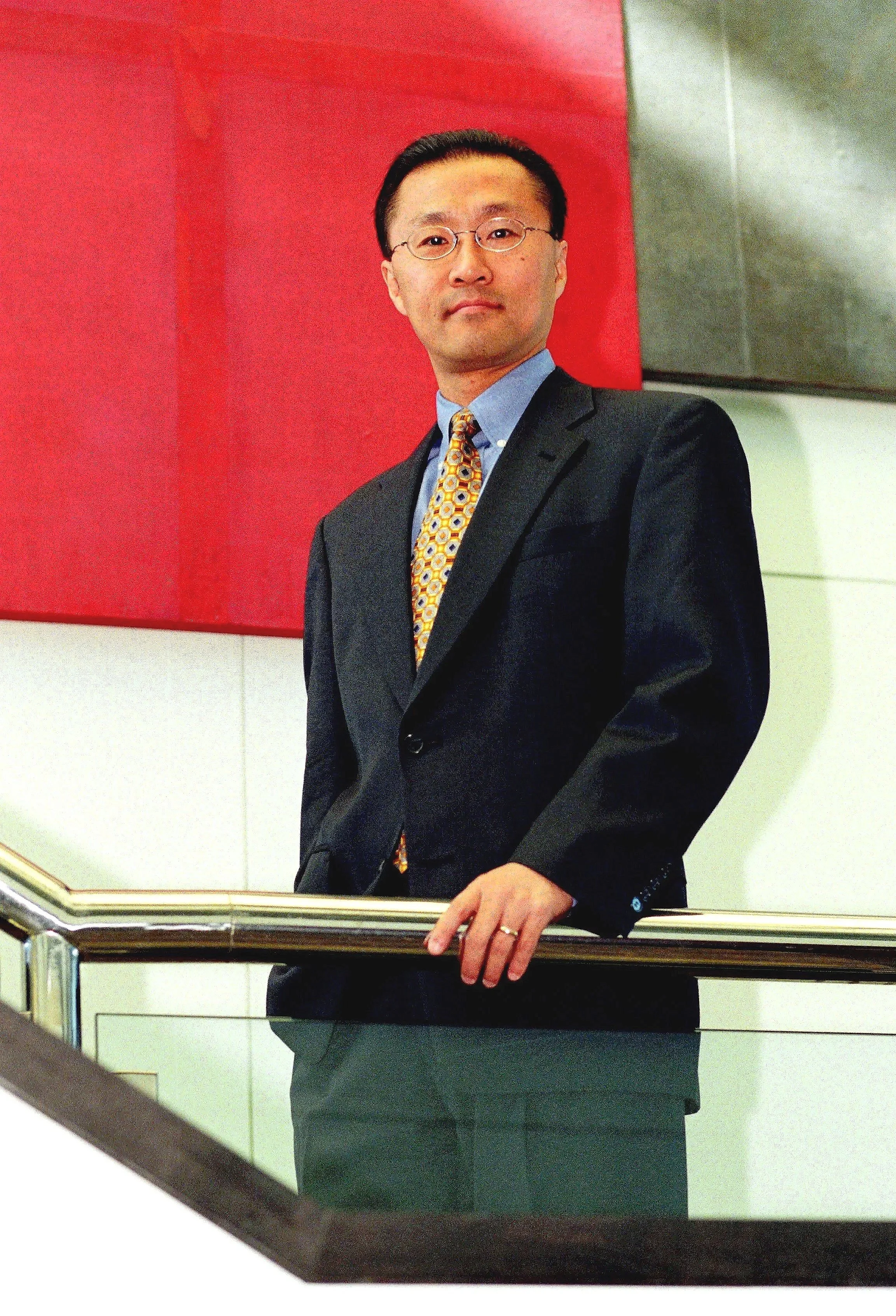 General Counsel for IKON Business Solutions wearing glasses, a dark suit, a blue shirt, and a colorful tie, standing behind a glass railing with a red wall in the background.