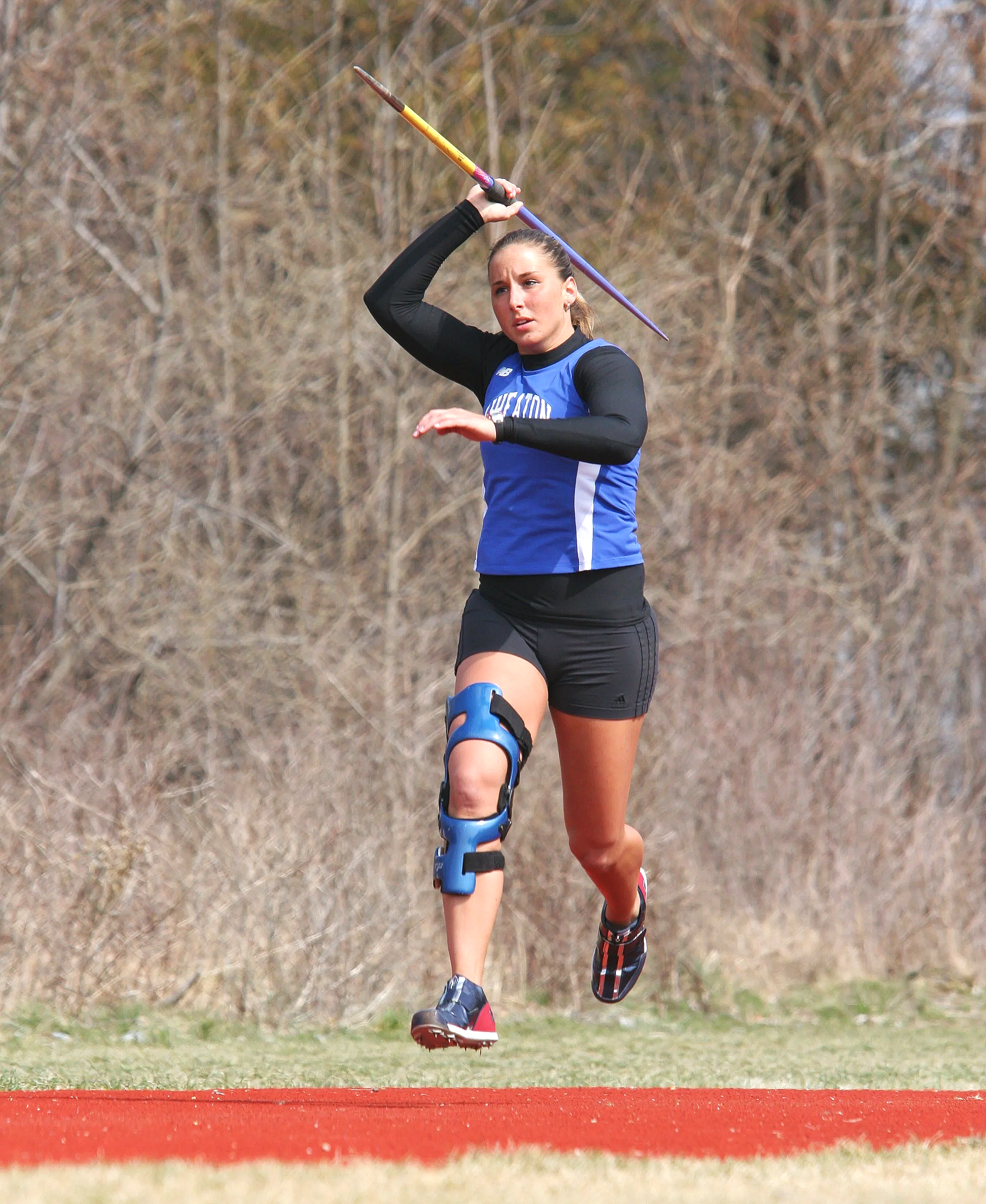 A female athlete in a blue sports uniform is running on a track with a pole in her right hand, and she is wearing a prosthetic on her right leg. She appears to be in a competitive event outdoors.
