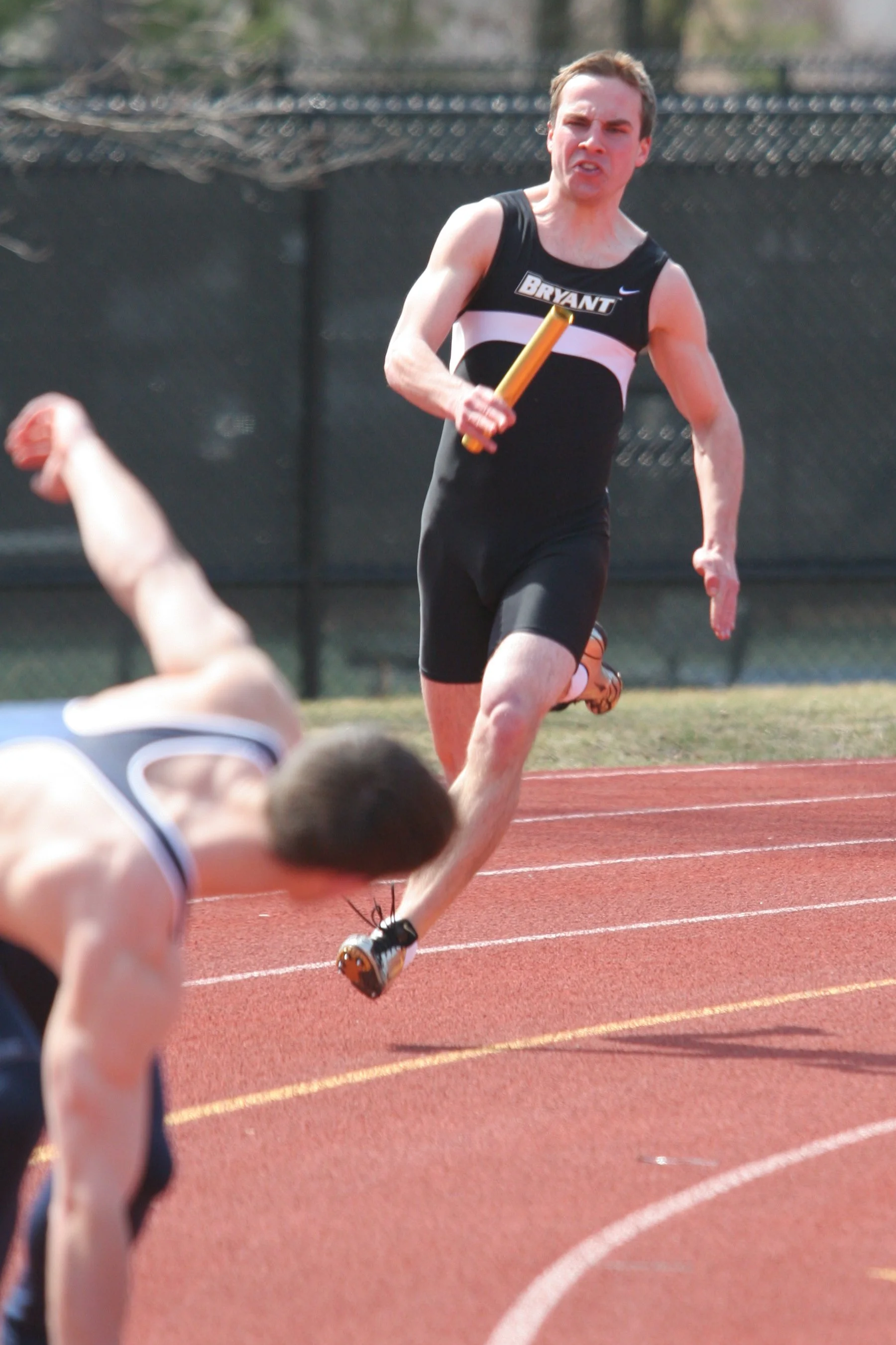 A male athlete in a black sports uniform running on a track, holding a relay baton, with another runner falling or lying on the ground nearby.