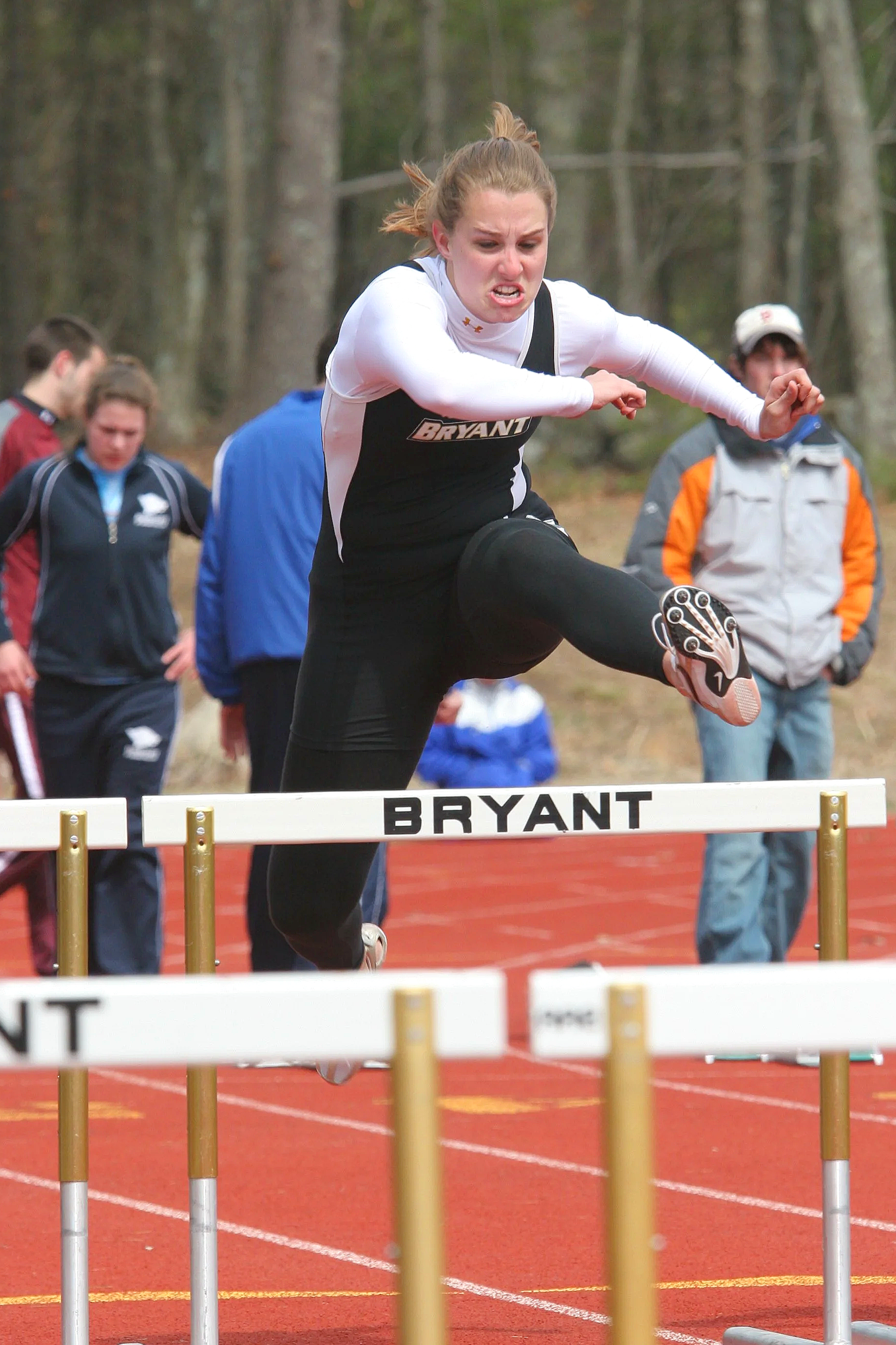 A woman in athletic clothing is in mid-air jumping over a hurdle on an outdoor track during a race, with people watching in the background.
