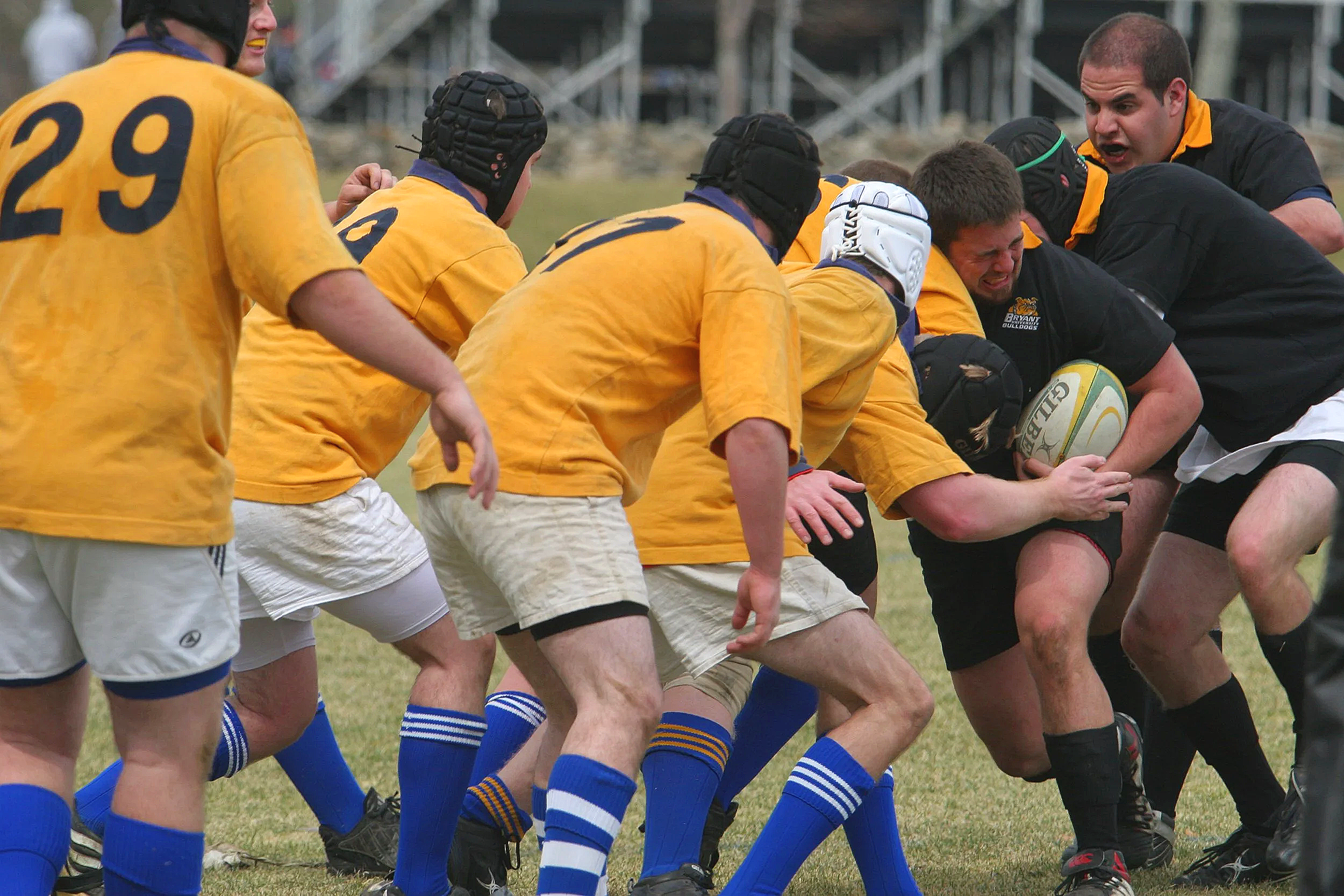 A group of rugby players in yellow jerseys and blue socks engaged in a scrum, with a coach or referee giving instructions nearby.