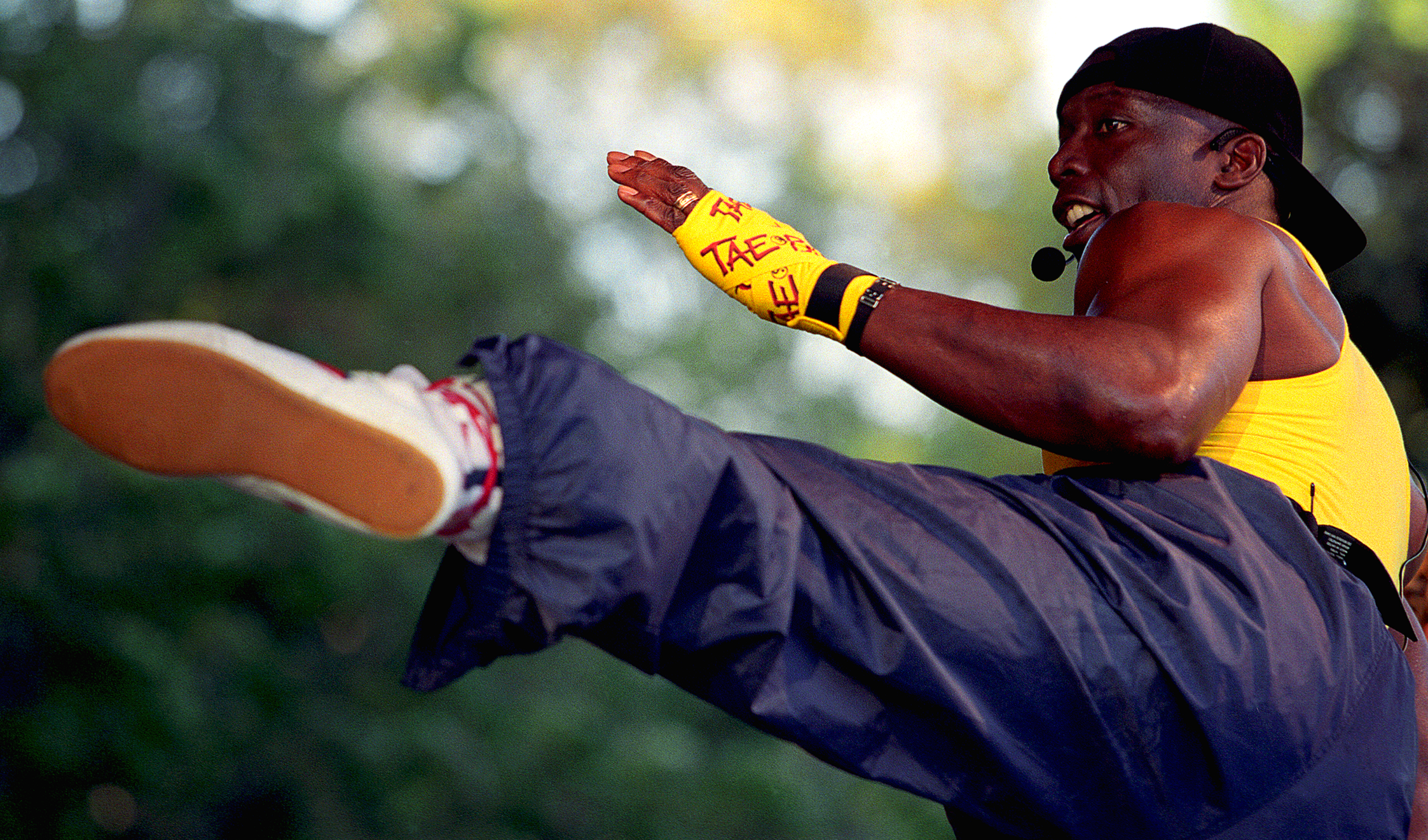 A man in a yellow tank top, black cap, and yellow taekwondo glove performing a high kick outdoors with a blurred green background.
