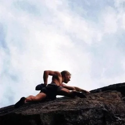 A man rock climbing outdoors on a steep, rugged cliff with a cloudy sky in the background.