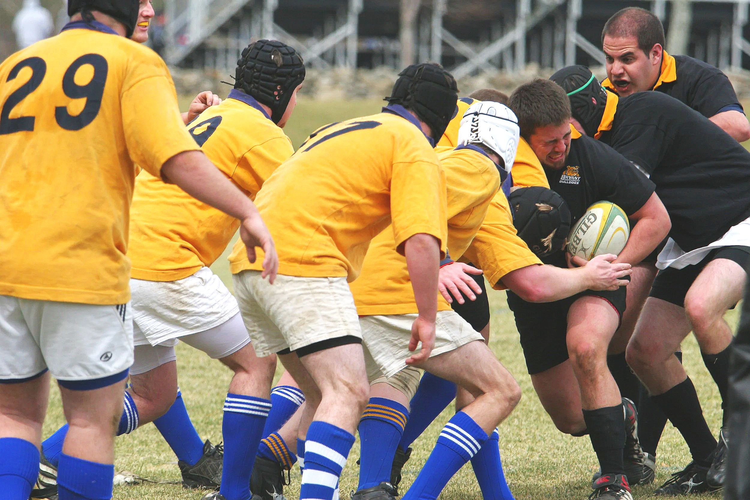 A group of rugby players in yellow jerseys and blue socks engaged in a scrum, with a coach or referee giving instructions nearby.