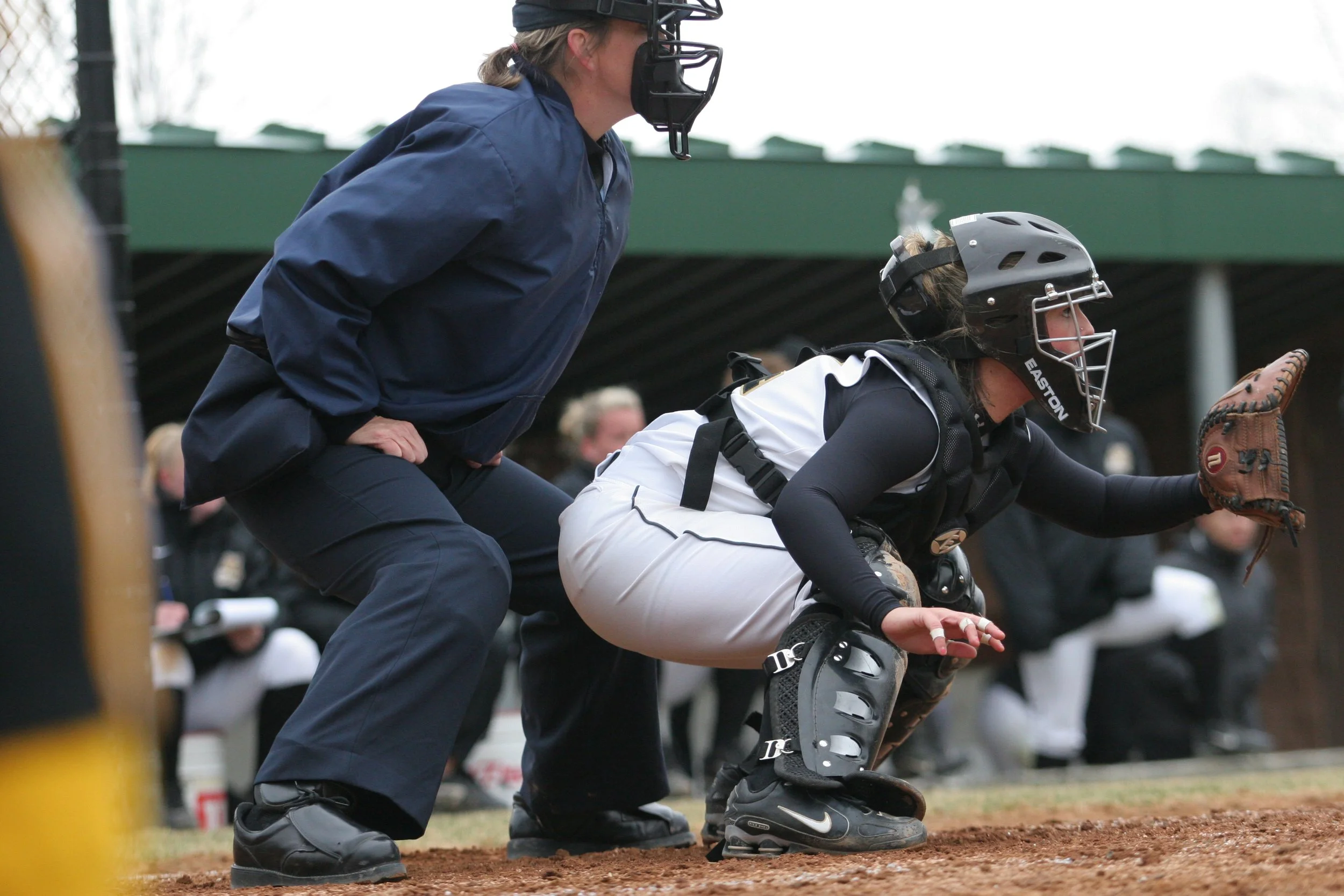 A female baseball catcher in protective gear crouches behind home plate, ready to catch a pitch, while a coach stands close behind her on a baseball field.