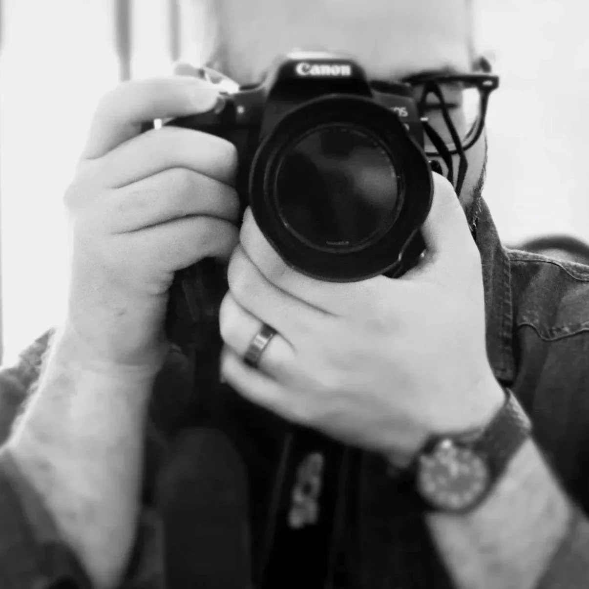 Black and white photo of a person taking a photo with a Canon camera, wearing a watch and a ring.