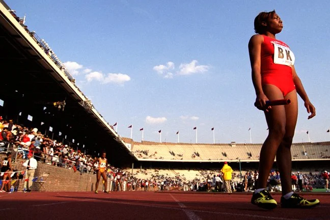 A female athlete standing on a track in a stadium, holding a relay baton, wearing a red athletic uniform with the letters B K, with other athletes and spectators in the background.