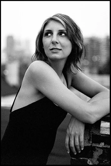 Black and white photo of a woman with shoulder-length hair, wearing a sleeveless top, sitting outdoors with a cityscape in the background.
