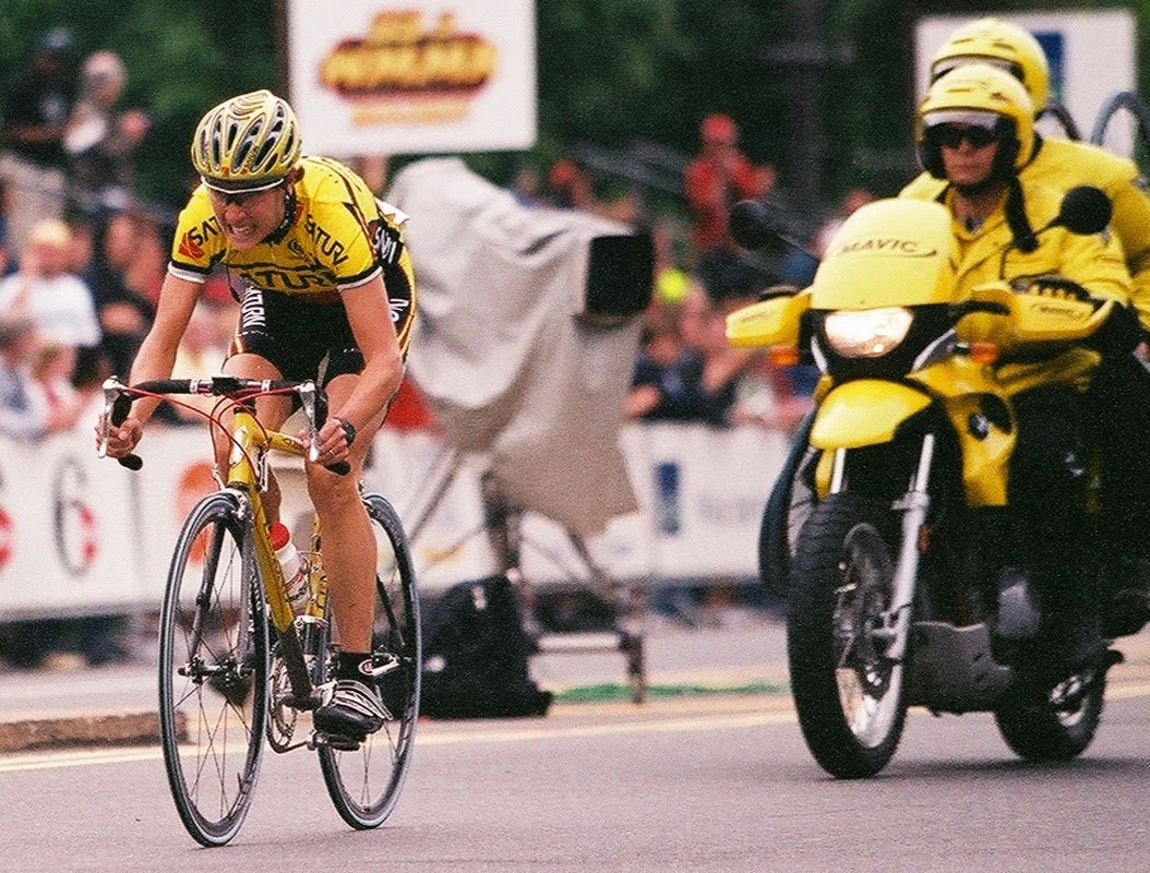 A cyclist wearing a yellow and black outfit and helmet is sprinting with a determined expression during a race, closely followed by motorcycle police officers also in yellow uniforms and helmets.