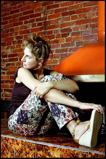 A woman with short, styled hair, wearing a dark purple top and colorful patterned pants, sits on a mosaic tiled surface with a brick wall background. She is sitting with her knees drawn up, looking to the side, with an orange stove or oven behind her